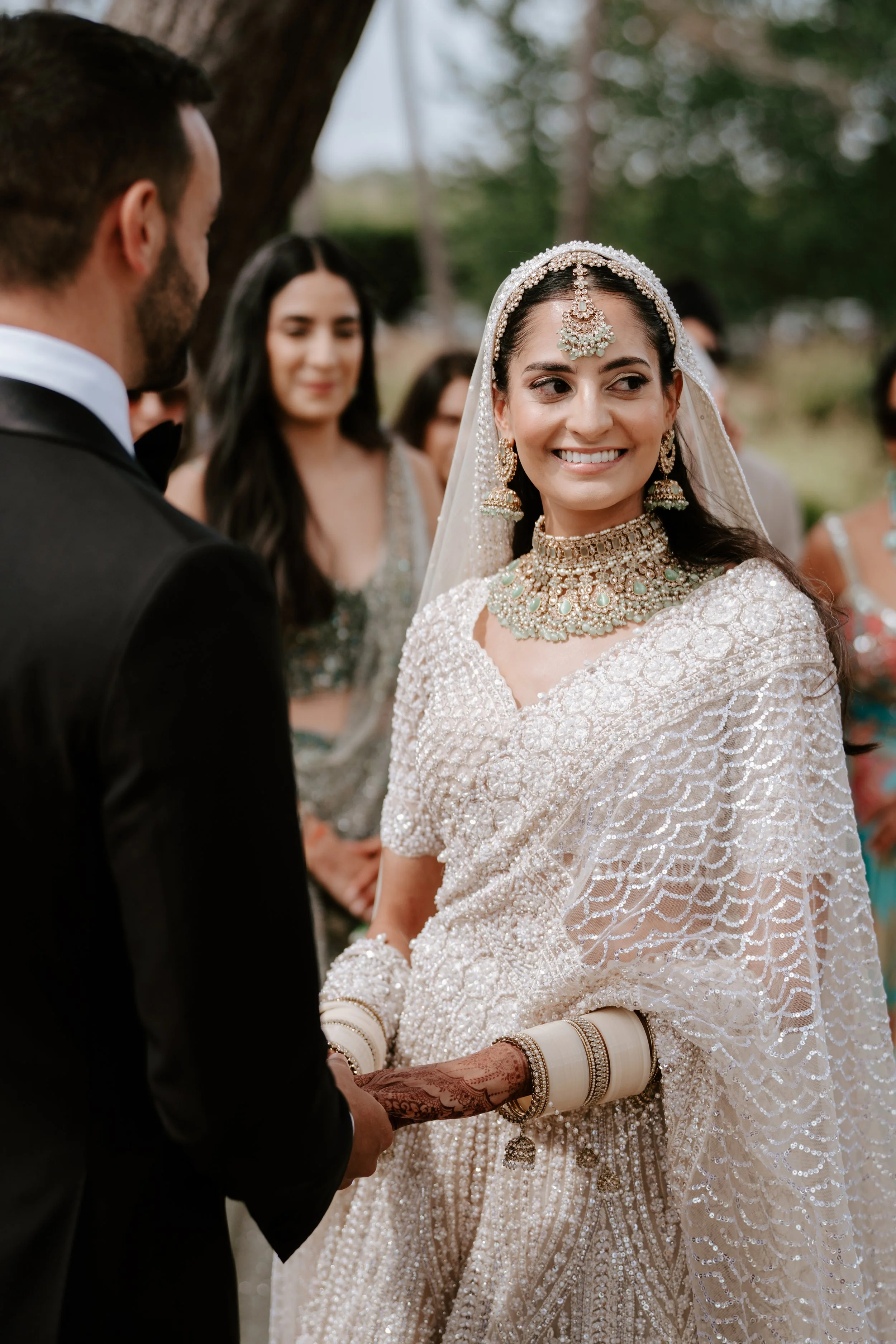 A bride smiling at her groom during a wedding ceremony outdoors, surrounded by guests and trees.