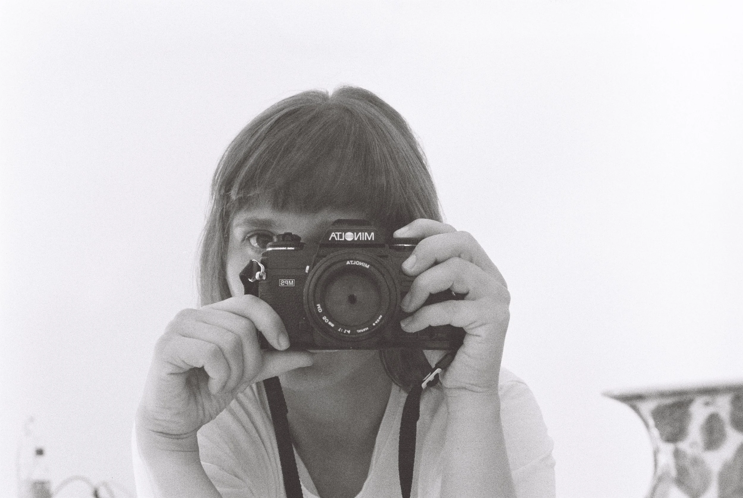 A person with brown hair holding a film camera, taking a selfie in front of a mirror.