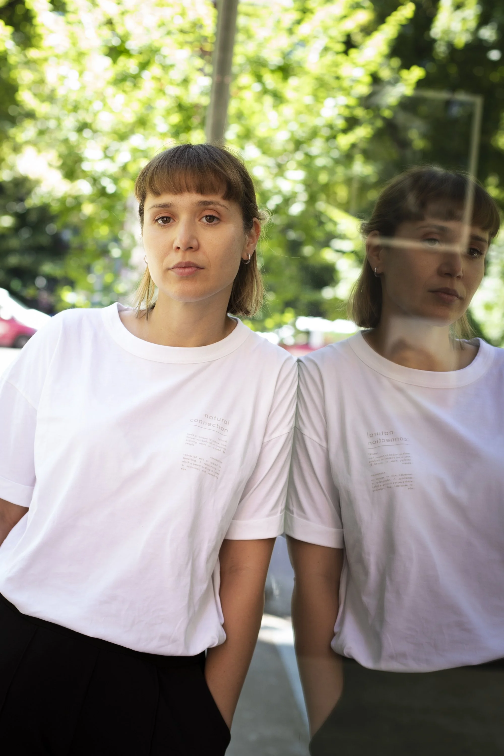A woman in a white T-shirt standing outdoors next to a reflective surface, creating a mirror image of her face and upper body, with green trees in the background.