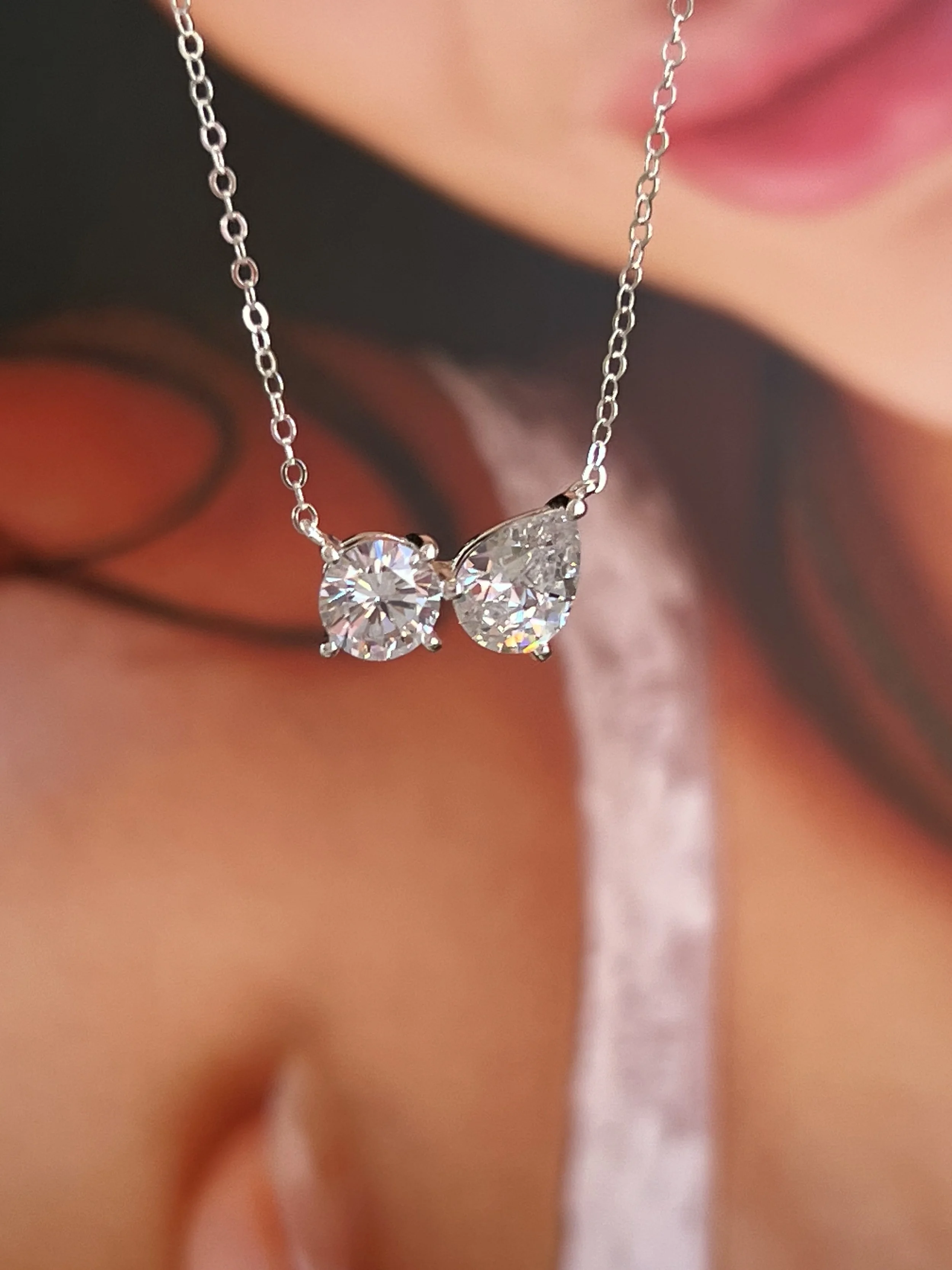 Close-up of a necklace with a butterfly-shaped pendant made of two large, clear, round-cut gemstones, held by a delicate silver chain. In the background, a person's lips and part of a face are slightly blurred.