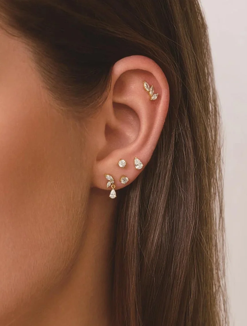 Close-up of a woman's ear with multiple gold and diamond earrings including a small ear cuff, a stud, a mini heart-shaped earring, and a drop earring.