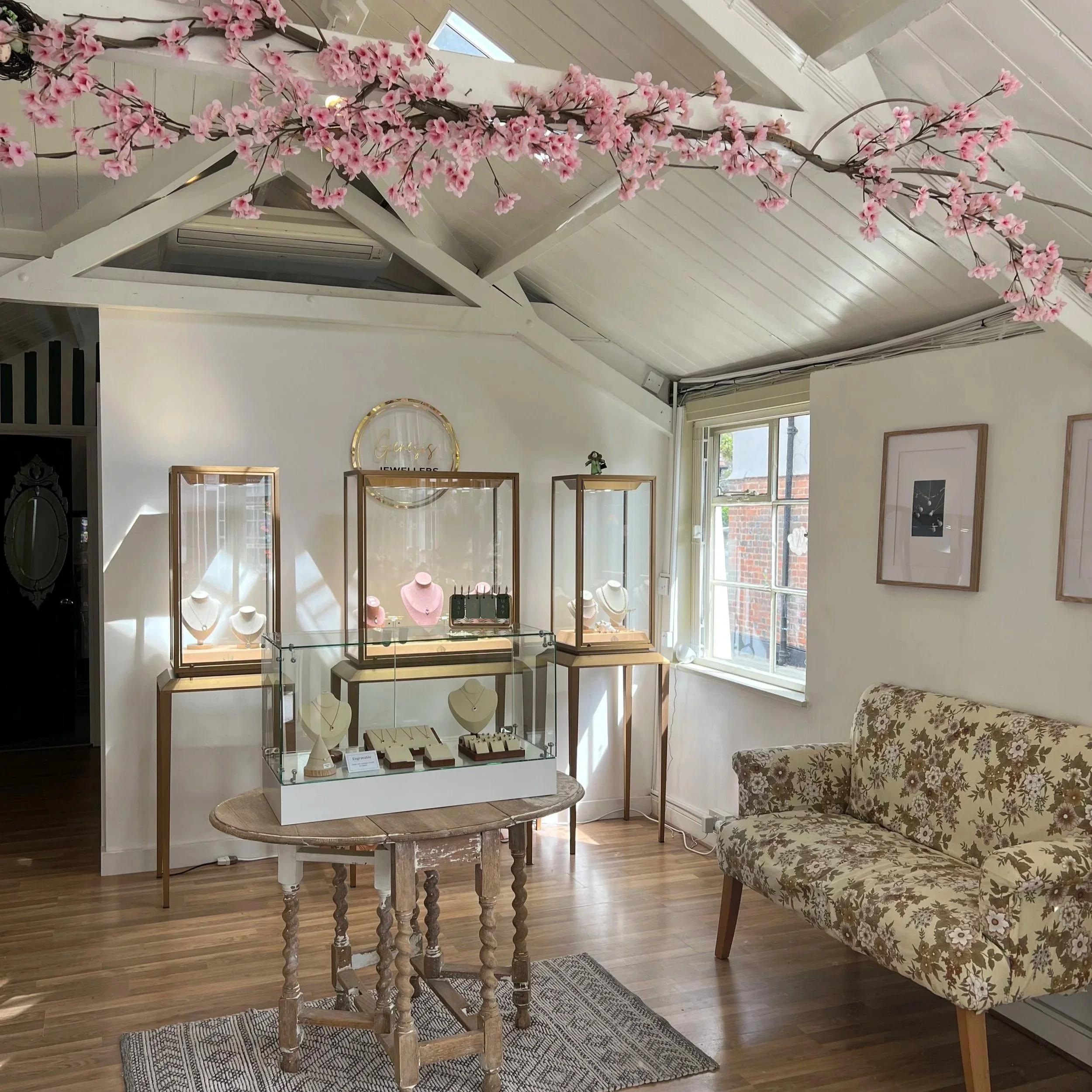 An interior shop display featuring a large pink cherry blossom branch hanging above glass jewelry cases with necklaces, situated in a bright room with large windows and wooden flooring.