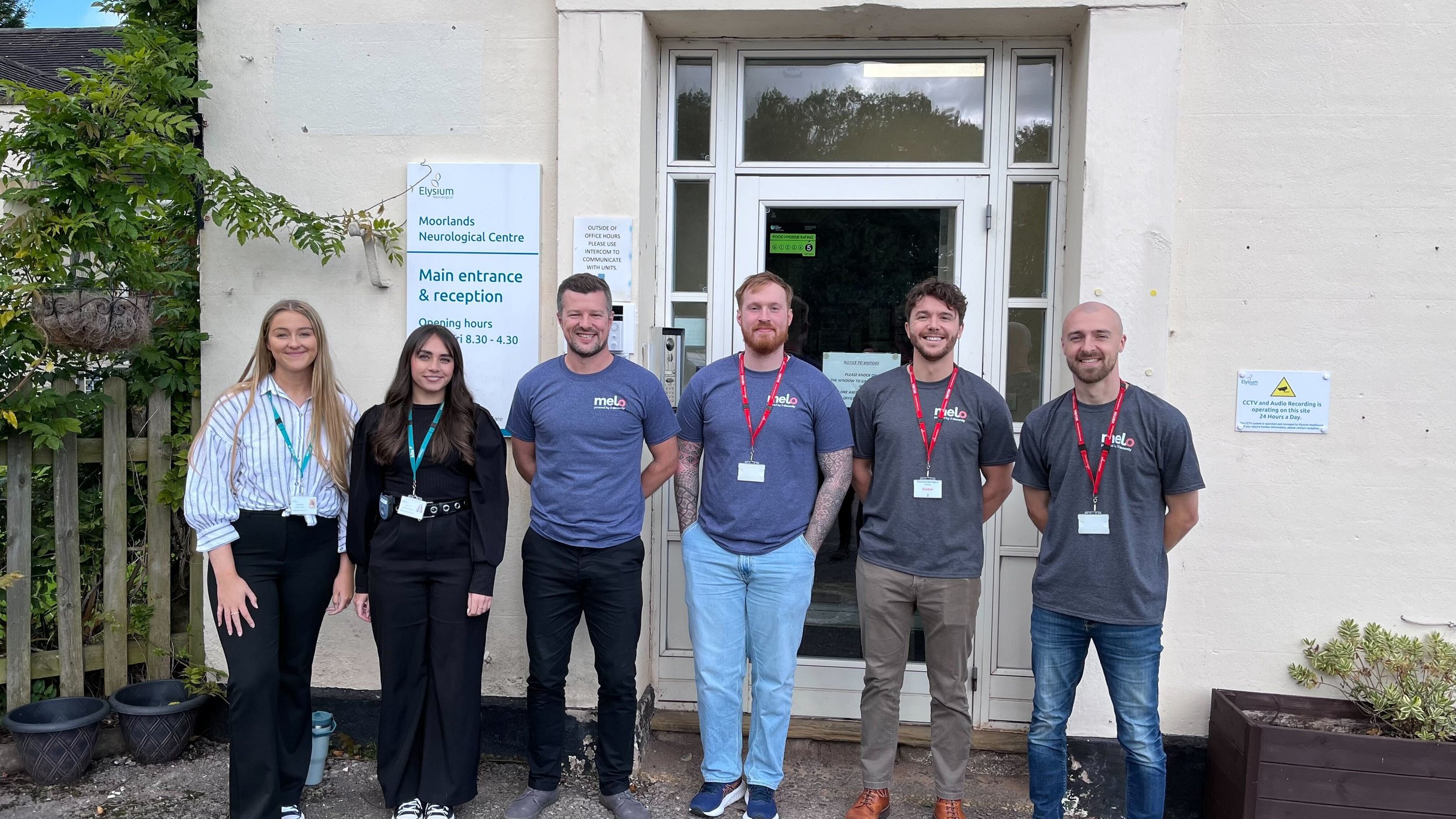 Group of six people standing outside the Moorlands Neurological Centre, in front of the main entrance with a sign showing opening hours.