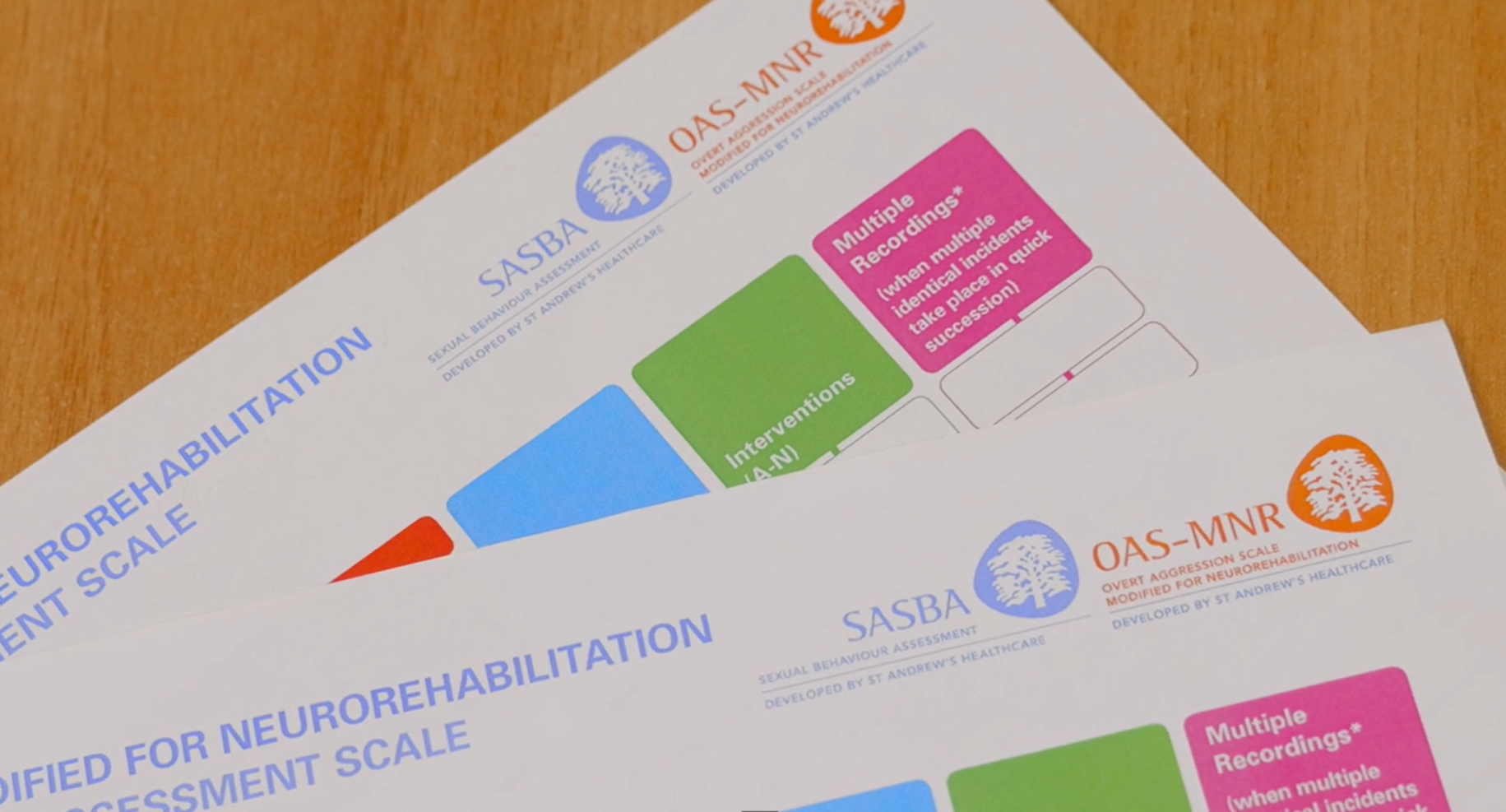 Two sheets of paper on a wooden surface, one partially visible with a color-coded chart and text about neurorehabilitation assessment scale, SET awareness, and intervention guidelines.