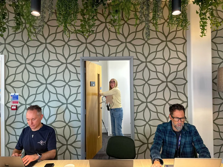 Two team members at their laptops while a colleague looks in from a doorway