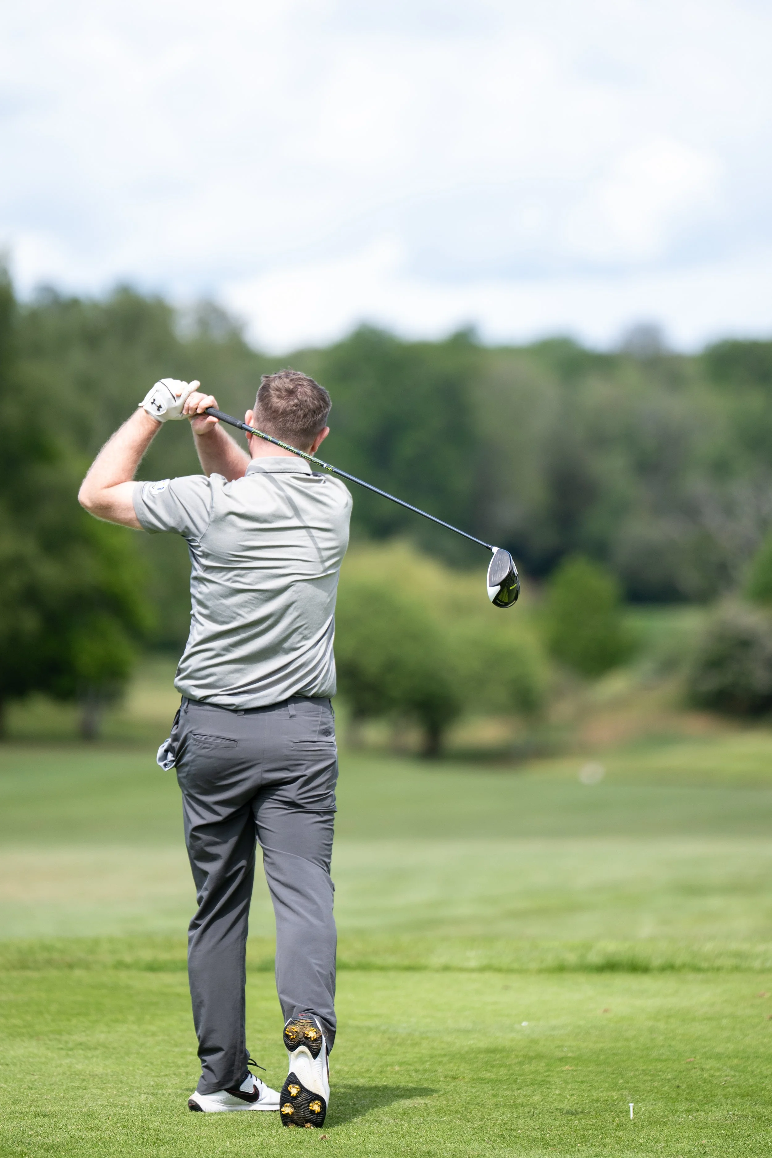 A man dressed in golf attire is swinging a golf club on a lush green golf course with trees in the background under a cloudy sky.