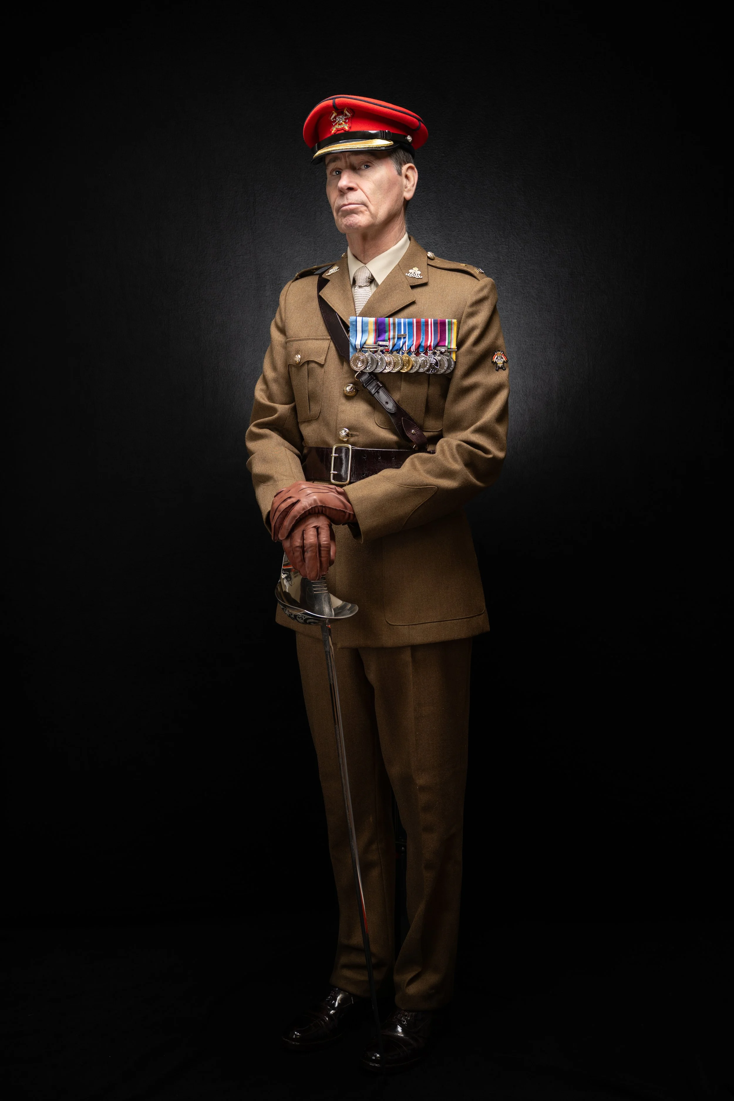 A man dressed in a military uniform with numerous medals, wearing a red and black peaked cap, holding a ceremonial sword, standing against a black background.