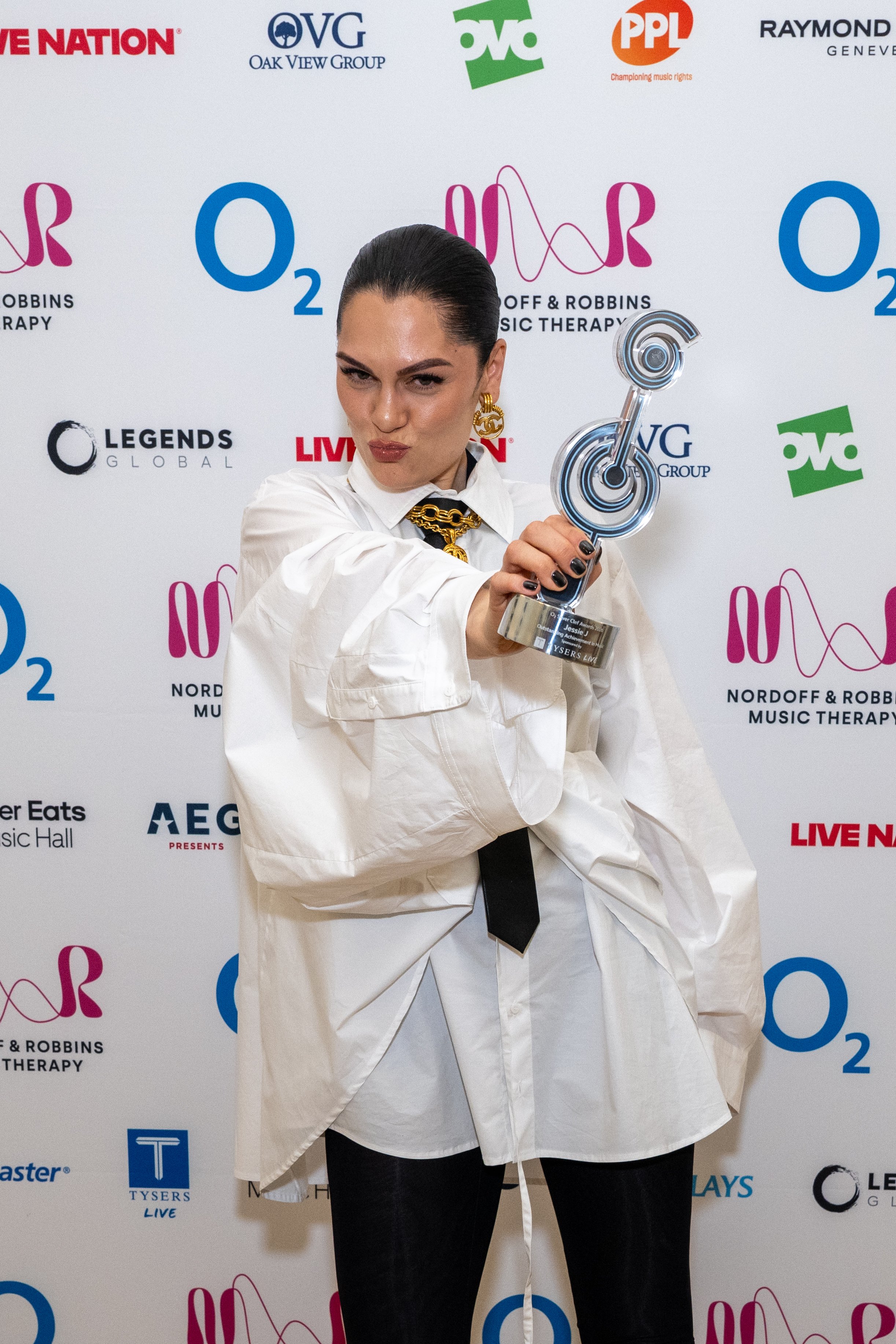 hair styled back, wearing a white oversized shirt, black pants, and a black tie, holding and pointing a silver trophy towards the camera, with a confident expression. The background features logos of O2, Nordoff & Robbins Music Therapy, and other sponsors.