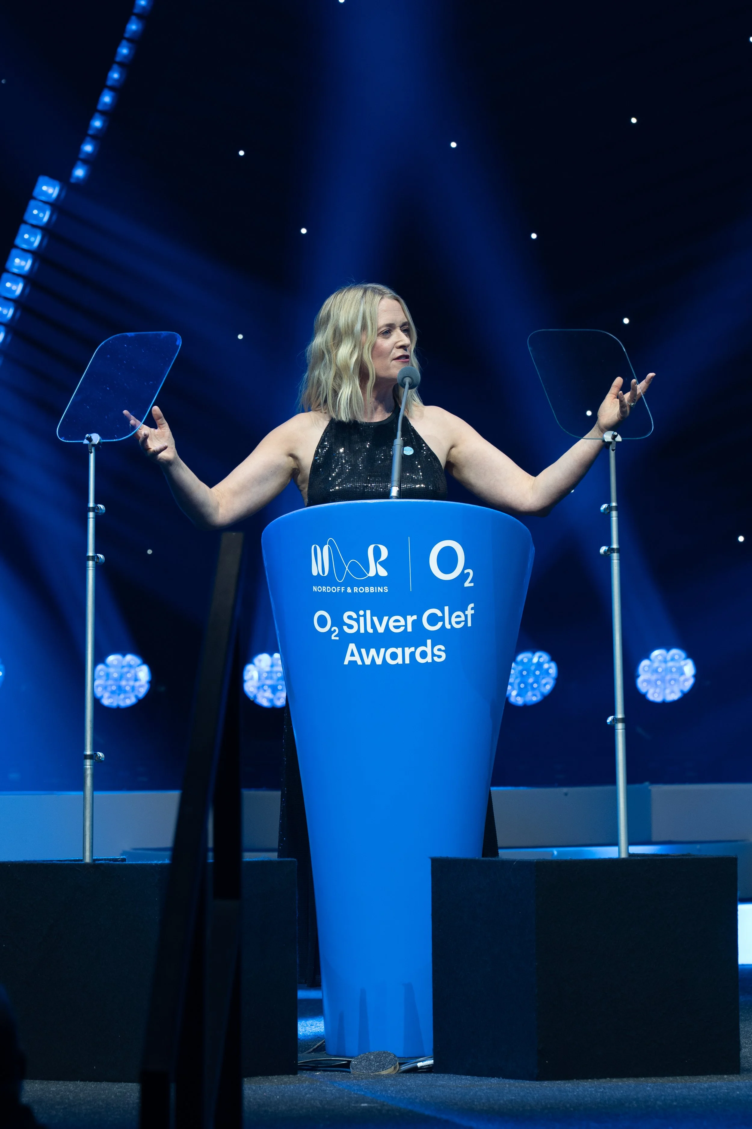 Edith Bowman standing behind a blue podium with logos for Nordoff & Robbins and O2, speaking into a microphone, with her arms raised and outstretched, at the O2 Silver Clef Awards event, with stage lights and a dark background.