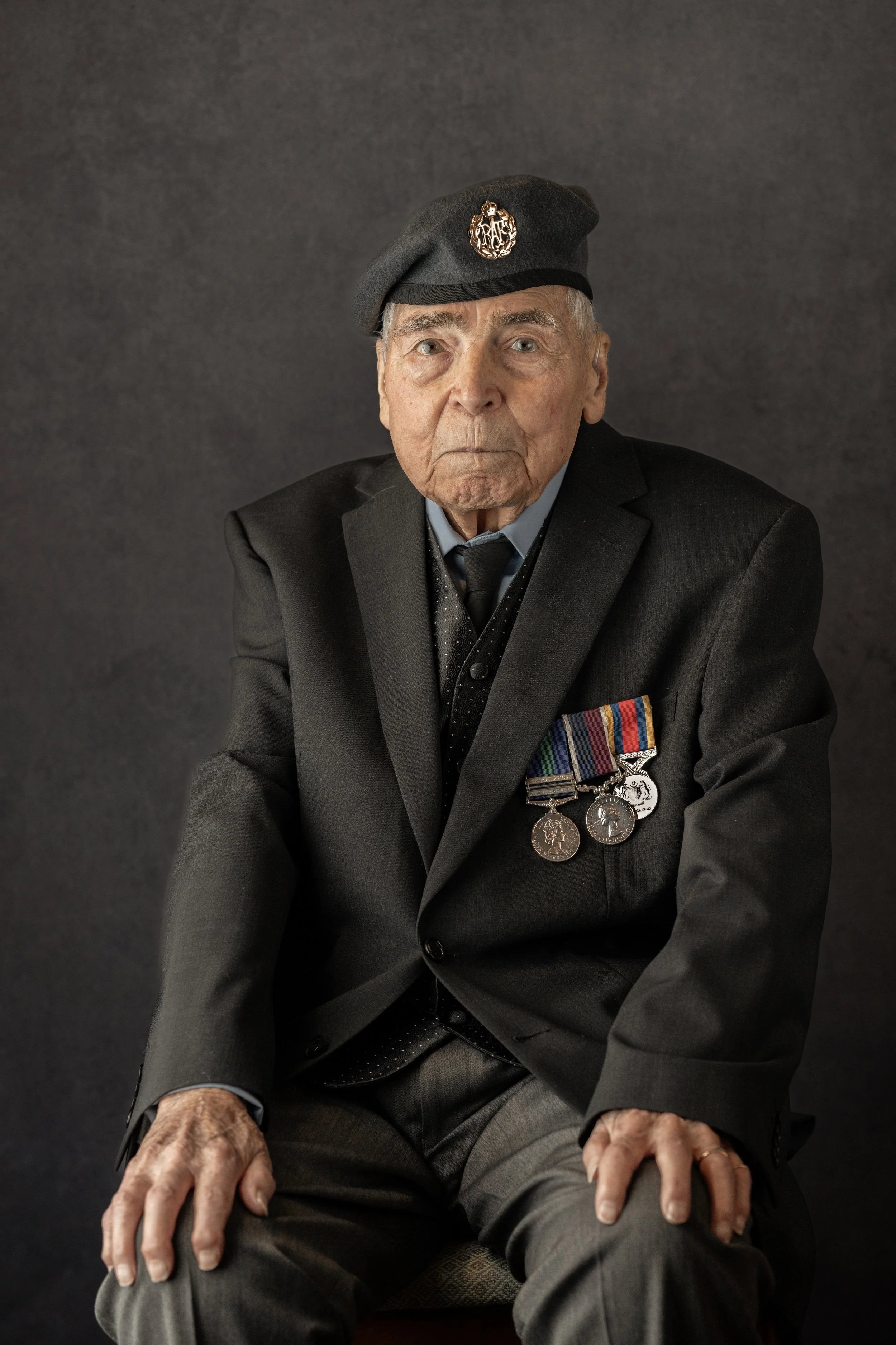 An elderly man in a dark suit with military medals and a beret with a badge, sitting against a dark background.