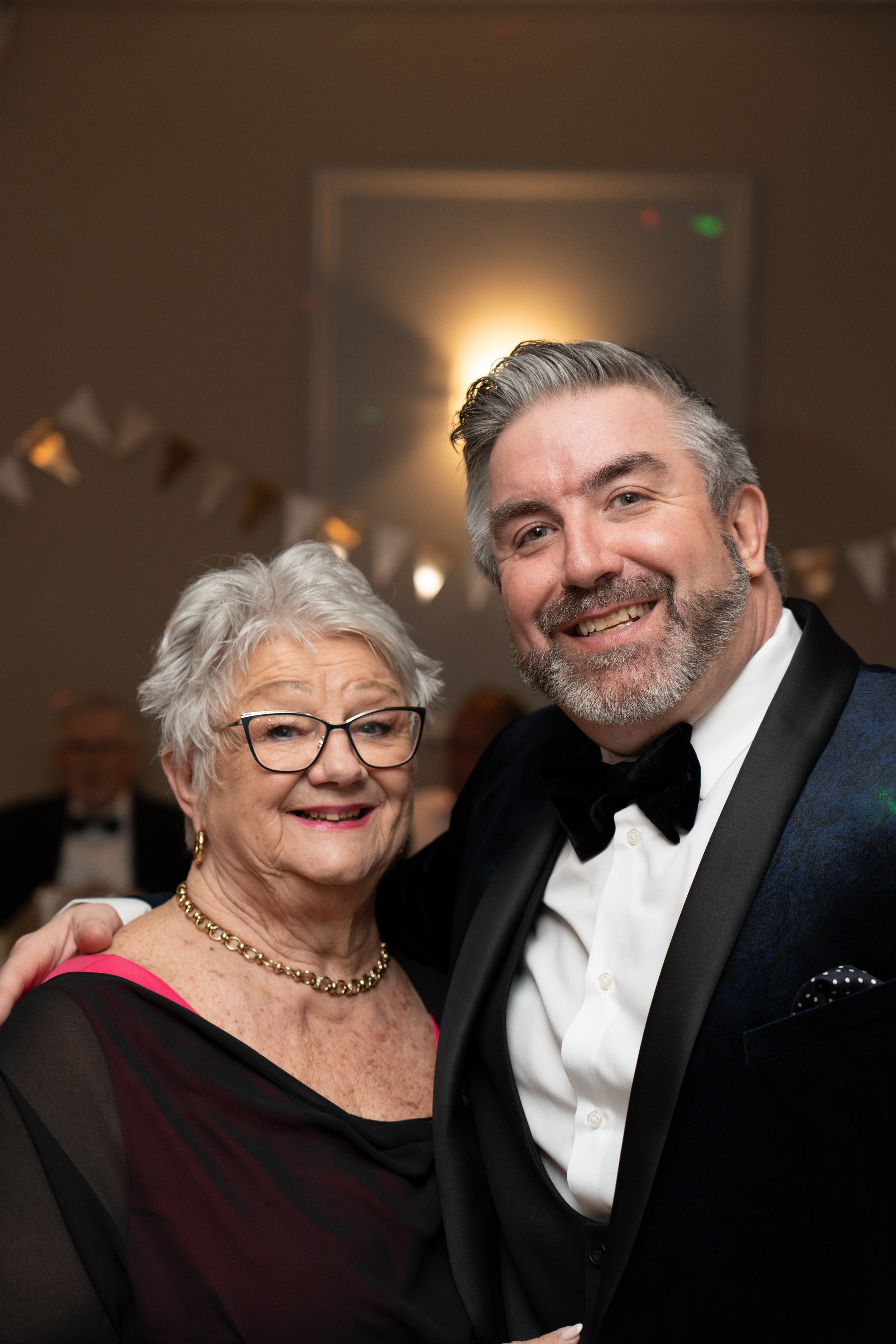 Two people smiling at a formal event, a woman with gray hair and glasses wearing a black and red dress with a gold necklace, and a man with gray hair and beard wearing a tuxedo with a bow tie.