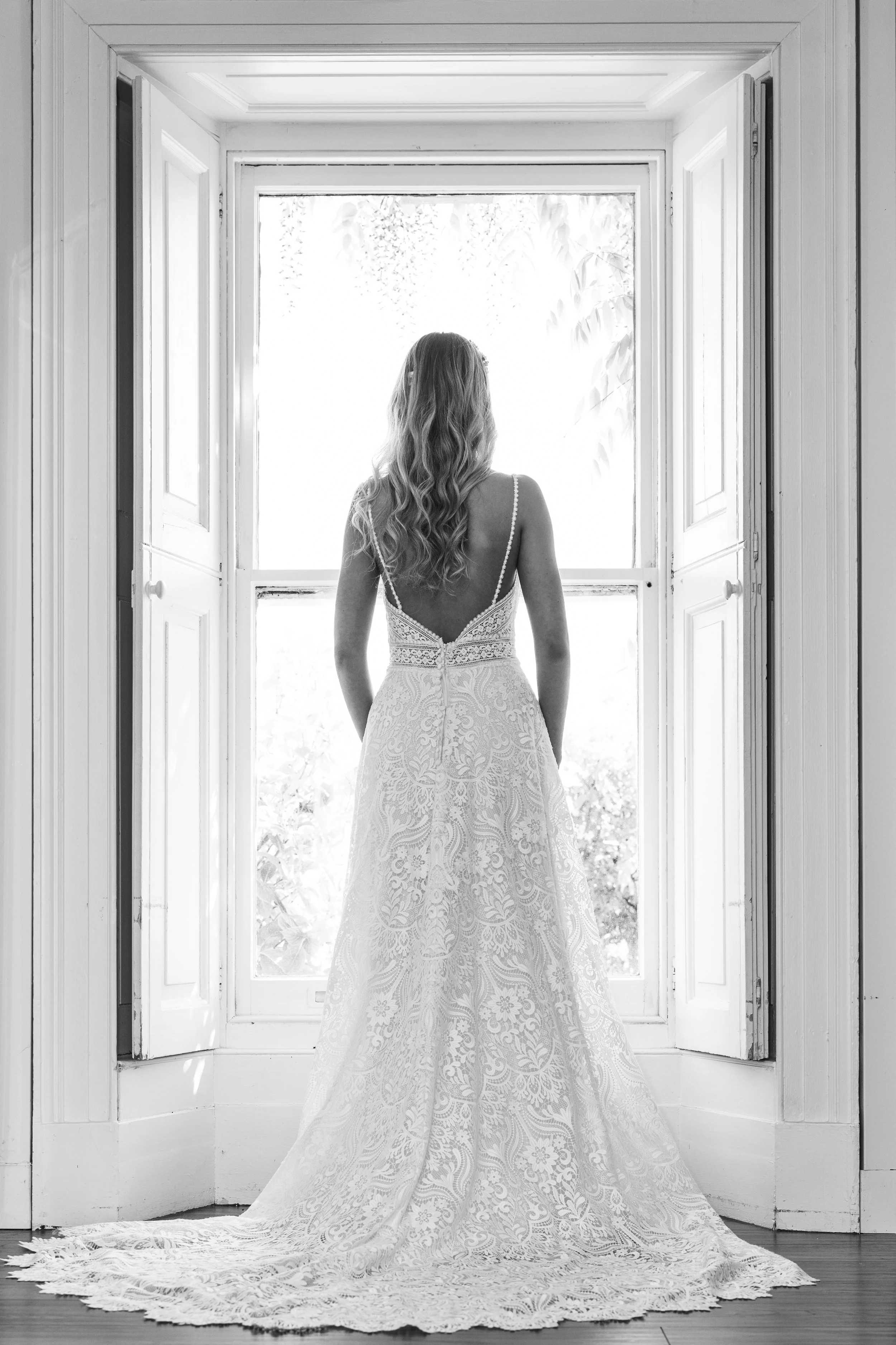 A woman in a lace wedding dress standing barefoot in front of a large window with open shutters, looking outside, in black and white.
