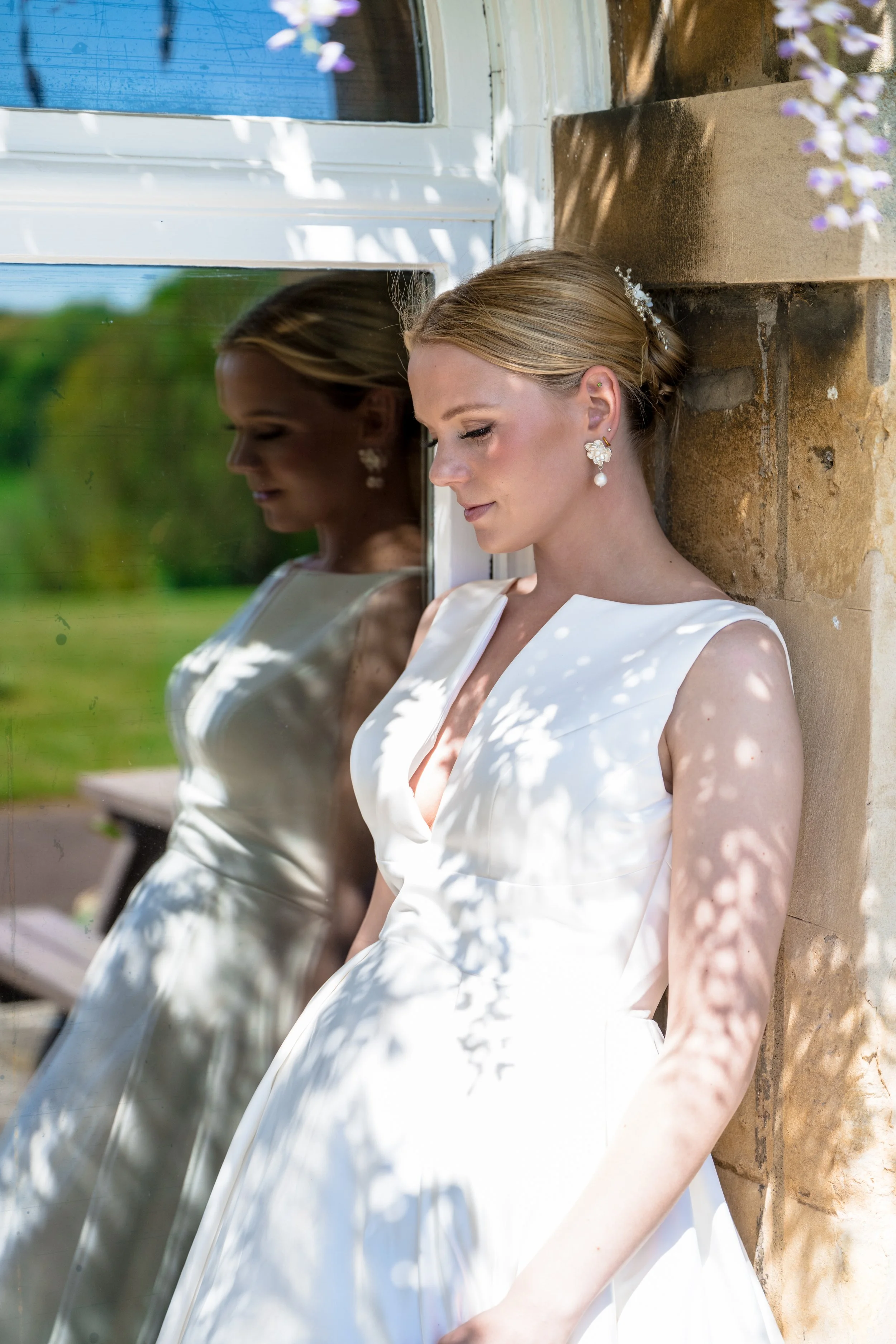 A bride in a white dress with pearl earrings and a decorative hair piece, rests her head against a window with her eyes closed, outdoors with green trees in the background.