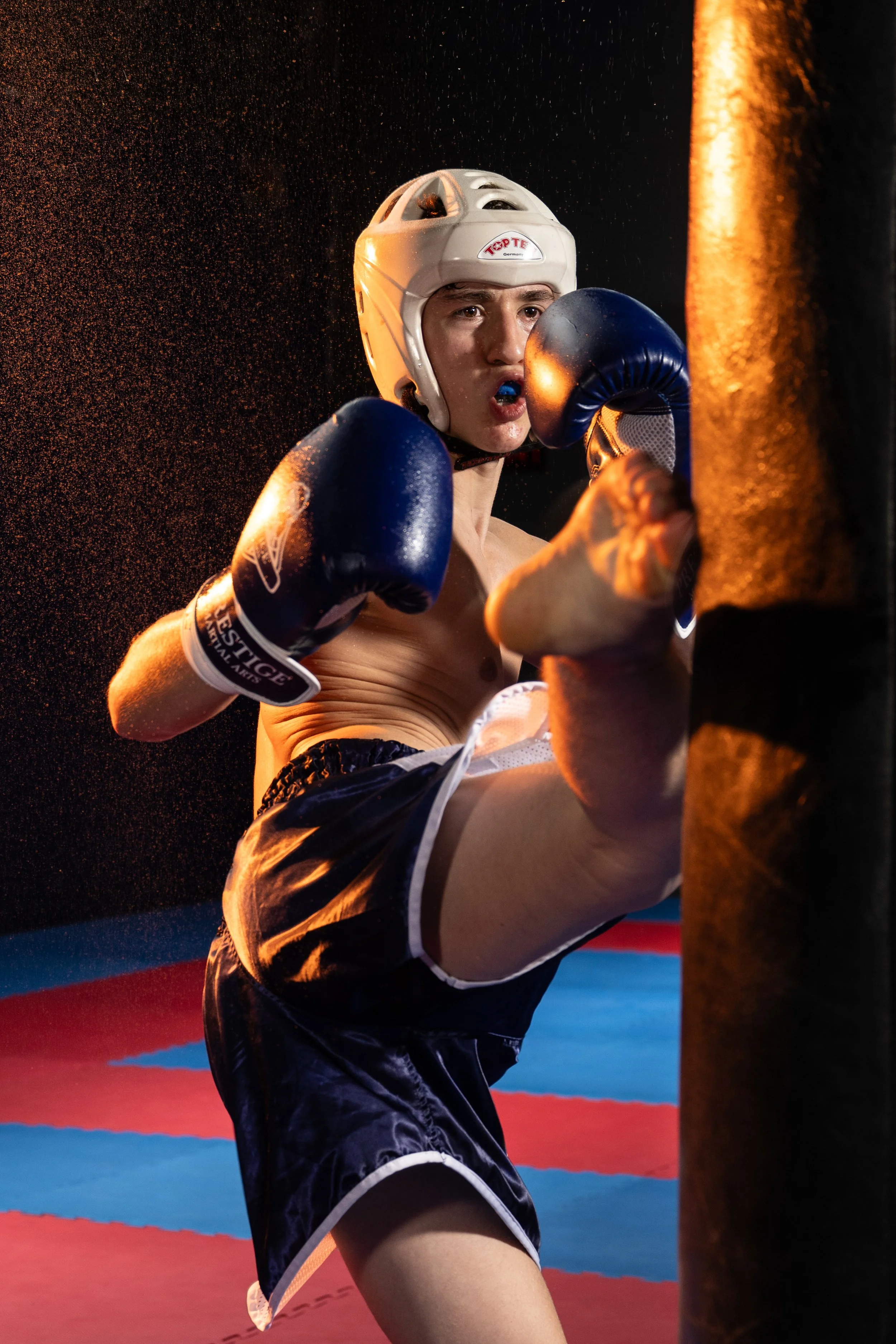 A male boxer wearing a white headgear and blue boxing gloves practicing punching a black punching bag in a boxing gym with red and blue mats.