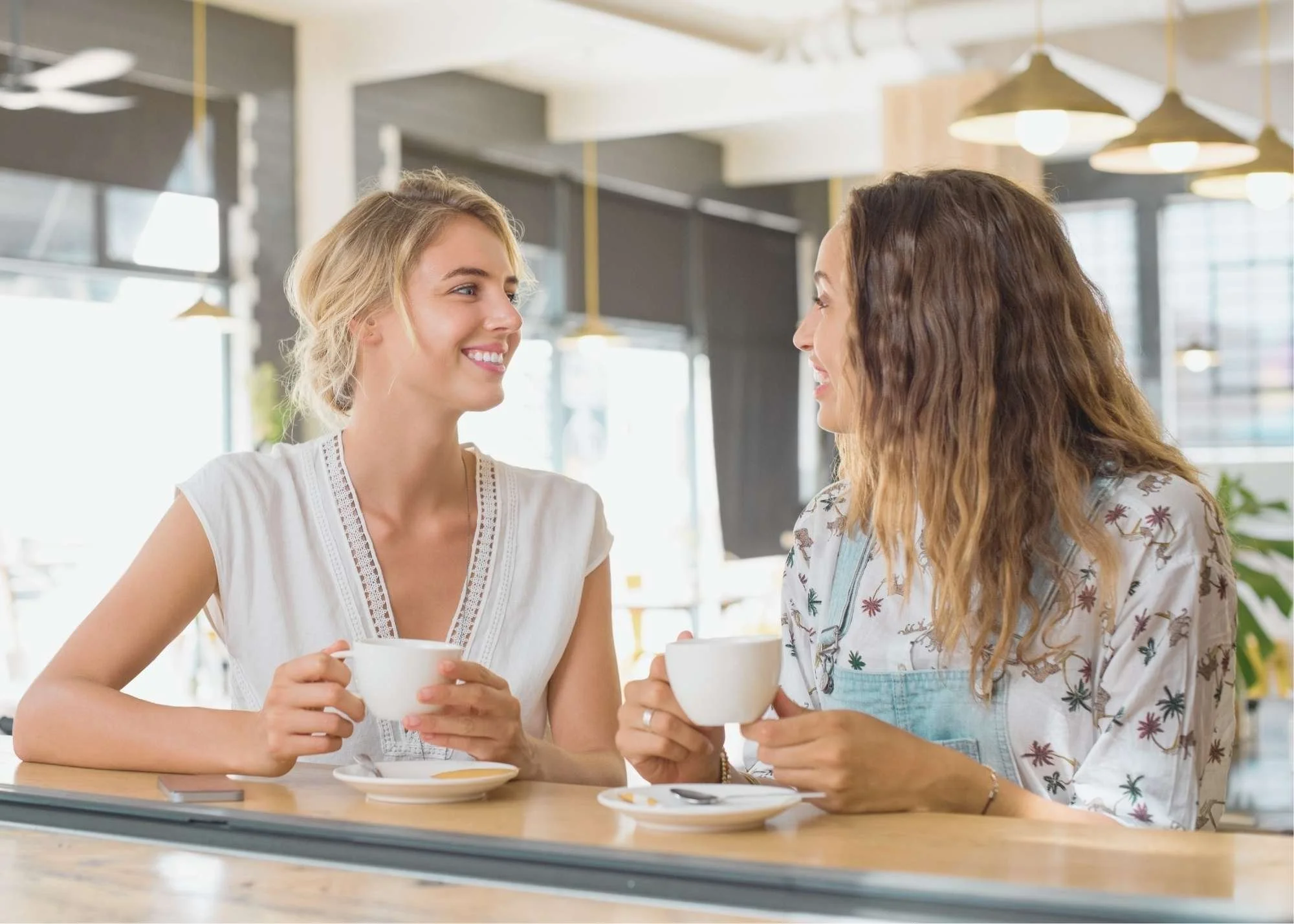 Two women sitting at a café, smiling and talking while holding white coffee cups
