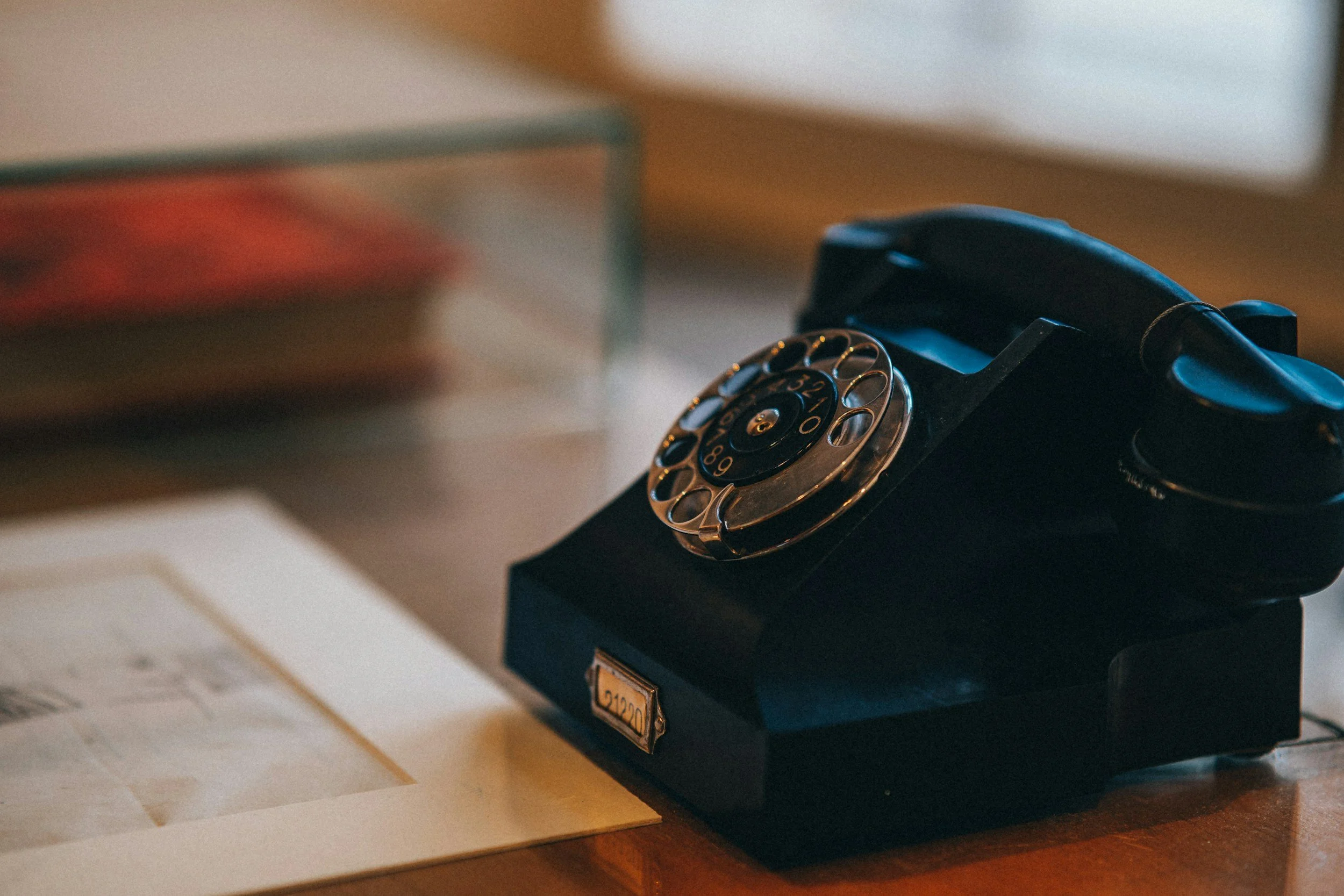 A vintage black rotary dial telephone with a gold-tone dial resting on a black stand, placed on a wooden surface next to scattered sheets of paper.