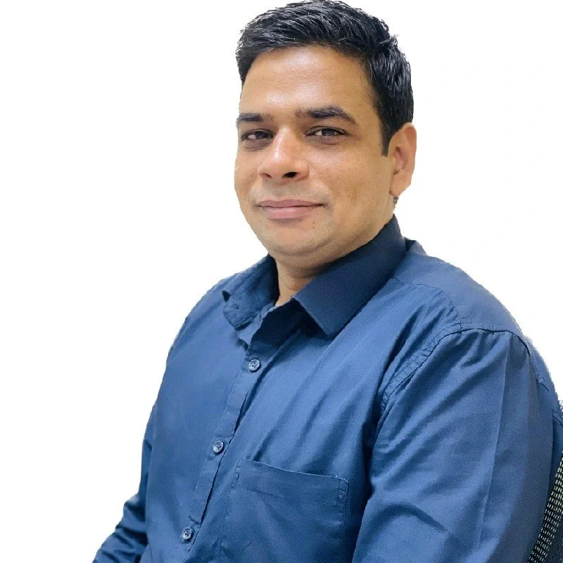 A man with dark hair and a light skin tone, wearing a blue button-up shirt, sitting and smiling at the camera against a plain white background.