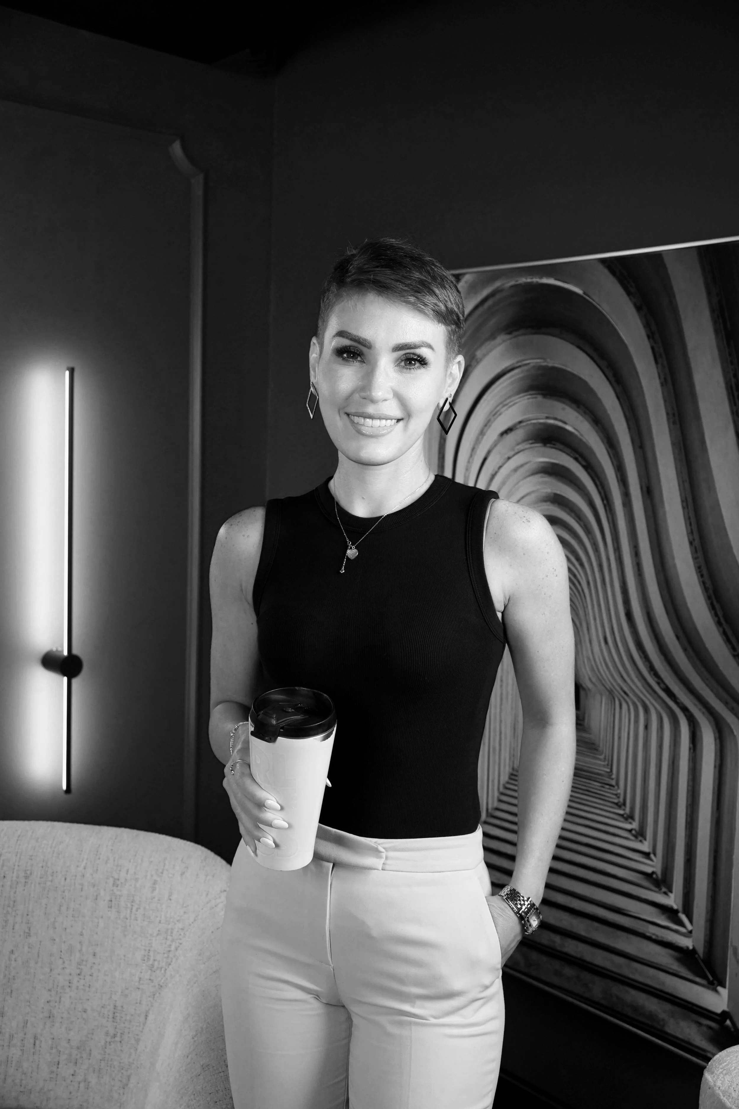 A woman with short hair wearing a sleeveless black top and white pants, holding a travel mug, and smiling in front of a colorful, patterned background.