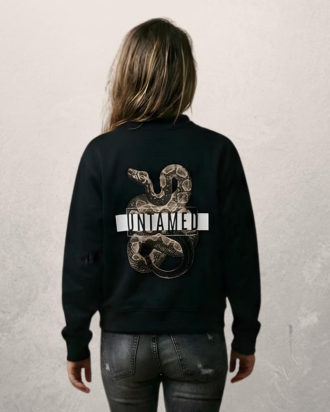 Person with long hair wearing our black Untamed Sweatshirt with a snake image and the word "UNNAMED" on the back, standing against a plain light-colored wall.