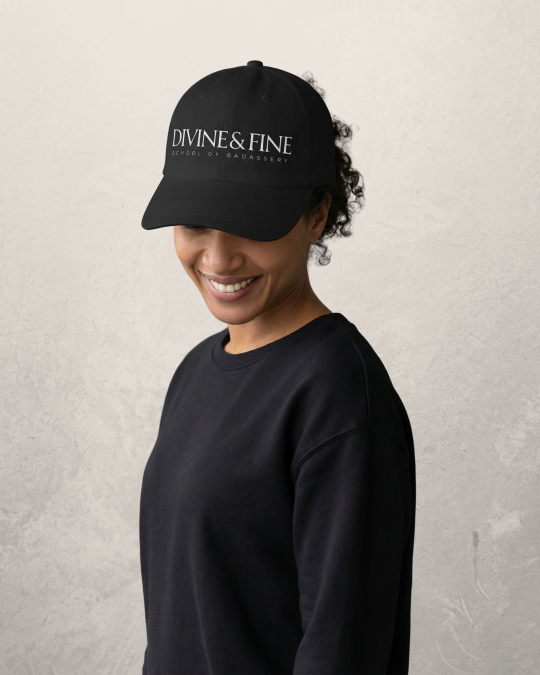 A woman in a black sweatshirt and black cap with white text that says, Divine & Fine, smiling with her eyes closed against a neutral background.