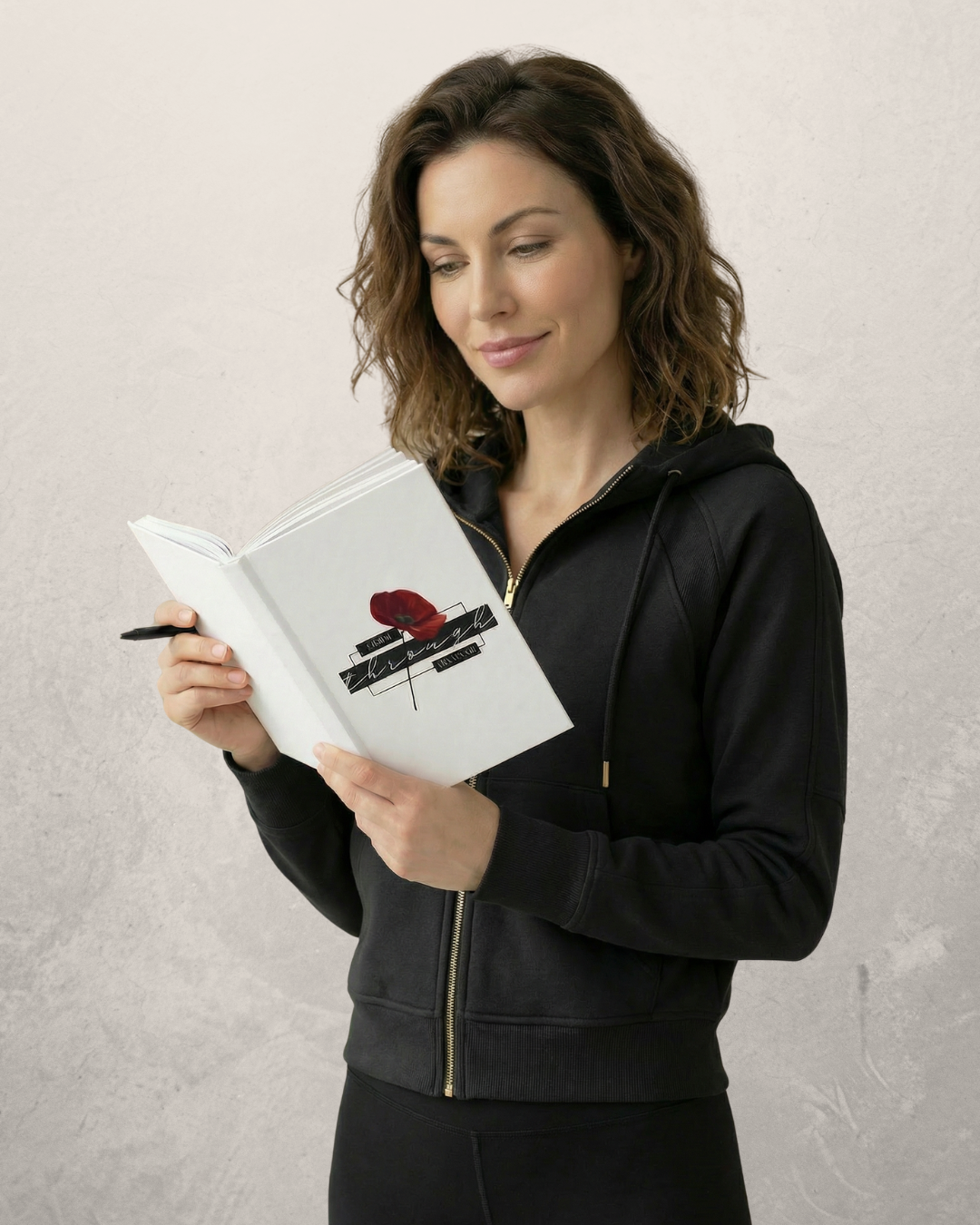 A woman with shoulder-length wavy brown hair reading our "I grow through the storm" journal with a poppy on the cover, wearing a black zip-up hoodie against a plain neutral background.