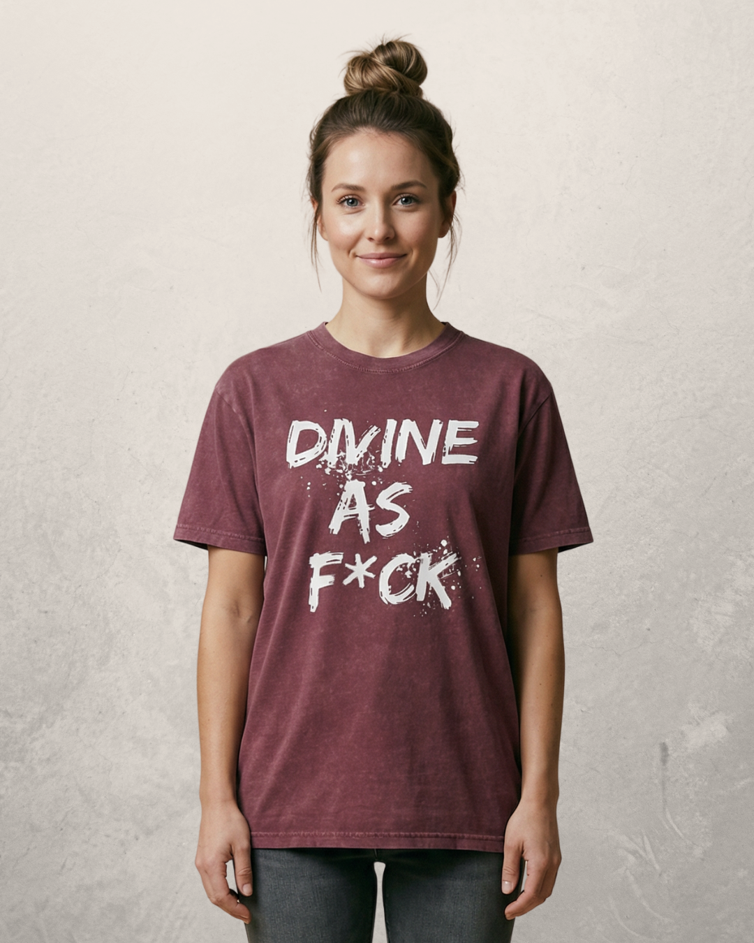 A young woman with brown hair in a bun wearing a maroon T-shirt that says "Divine as F*ck" standing against a beige wall.
