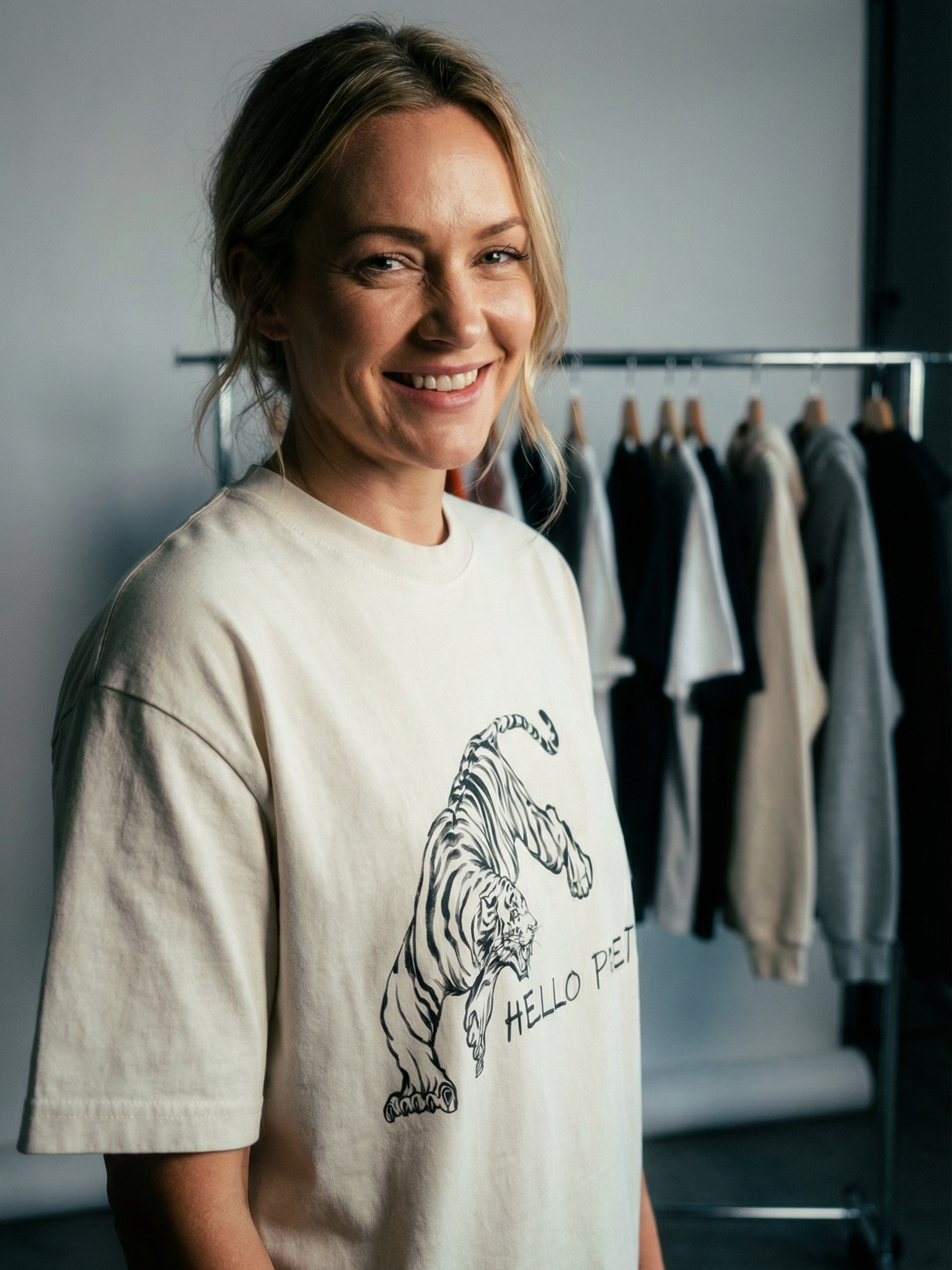Smiling woman in our cream T-shirt with a tiger illustration and the words "Hello Pretty" standing in front of a clothing rack with various shirts and shirts in the background.