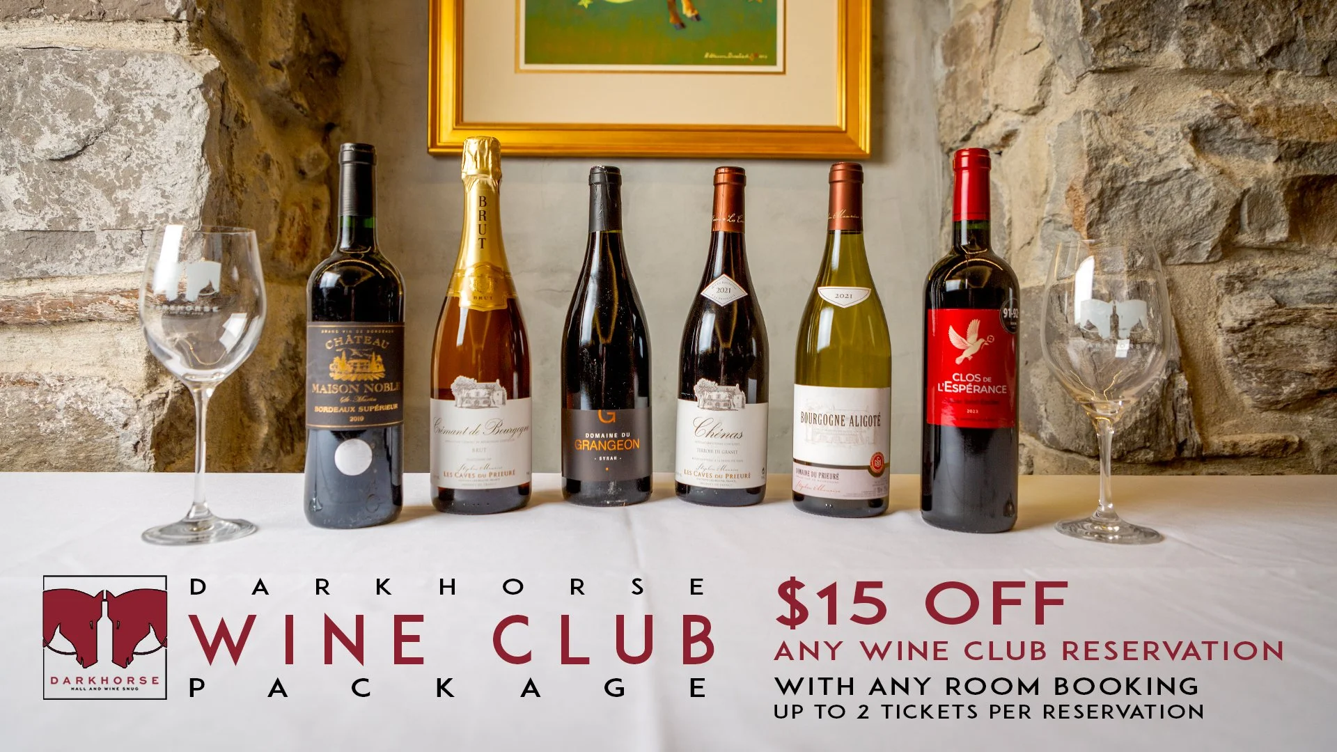 Six bottles of wine with two empty wine glasses on a white table, set against a stone wall and a framed picture. Overlay text advertises a Darkhorse Wine Club package with $15 off any reservation, up to two tickets.