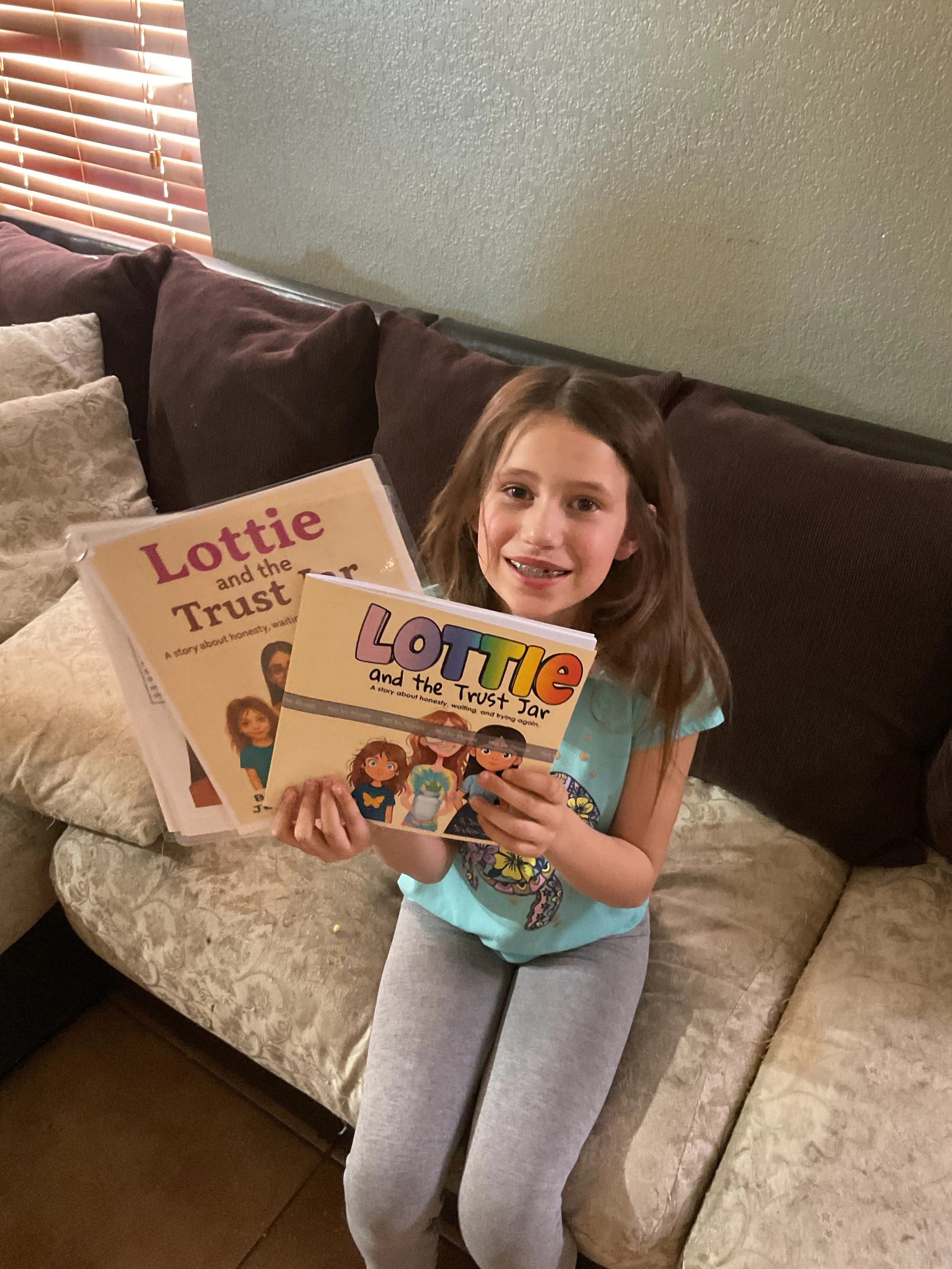 A young girl sitting on a beige patterned couch holding two children's books titled "Lottie and the Trust Jar" and "Lottie and the Trust Jar." She is smiling and has long, brown hair, wearing a light blue T-shirt with a graphic design and gray leggings.
