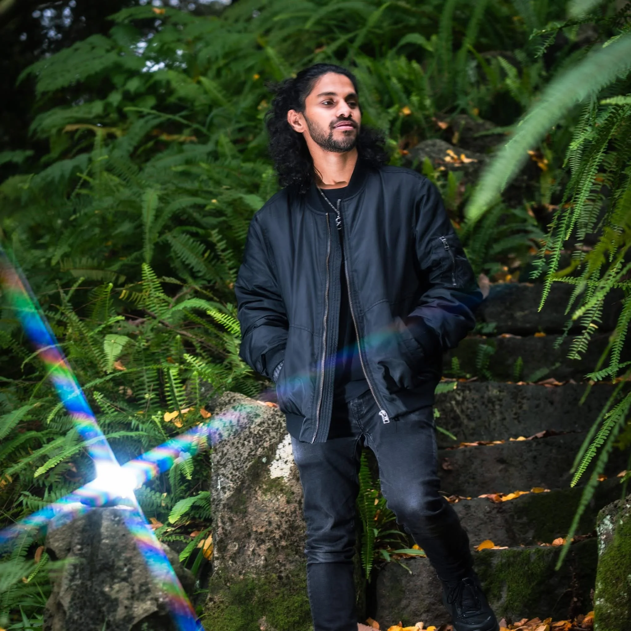 A man with long curly hair and a beard wearing a black jacket standing outdoors surrounded by lush green ferns and plants.