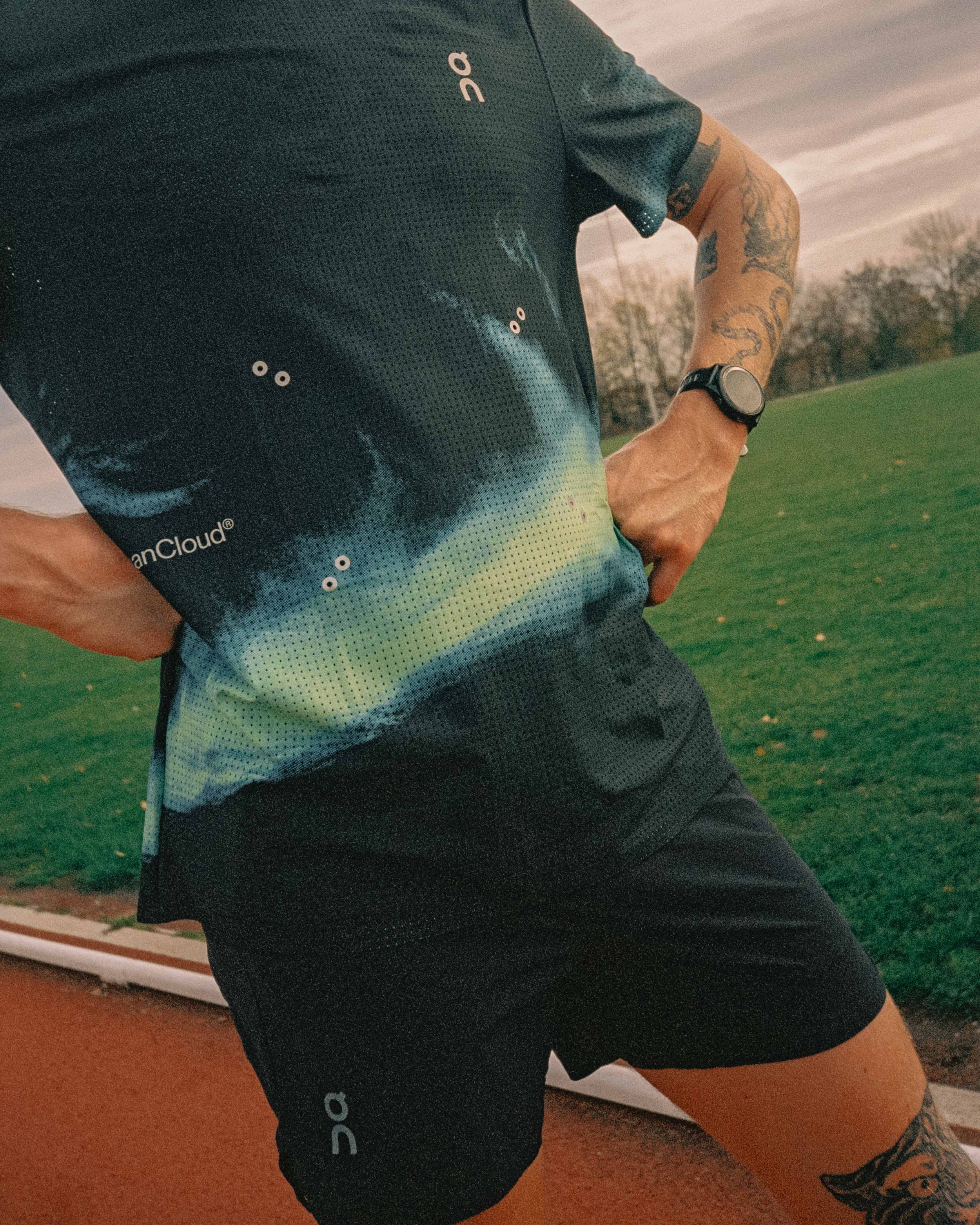 Close-up of a person wearing a black running shirt and shorts, standing outdoors on a track field with a grassy area and trees in the background during sunset.