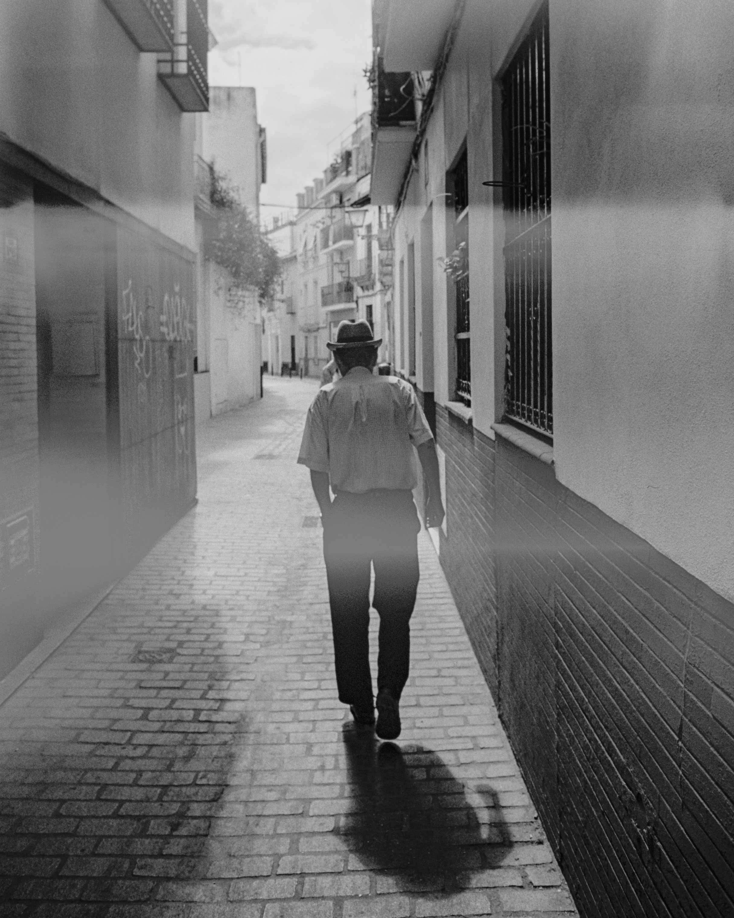 A man wearing a hat and a short-sleeve shirt walking down a narrow city street with brick pavement, carrying a notebook or folder in his left hand, with buildings and windows on either side.
