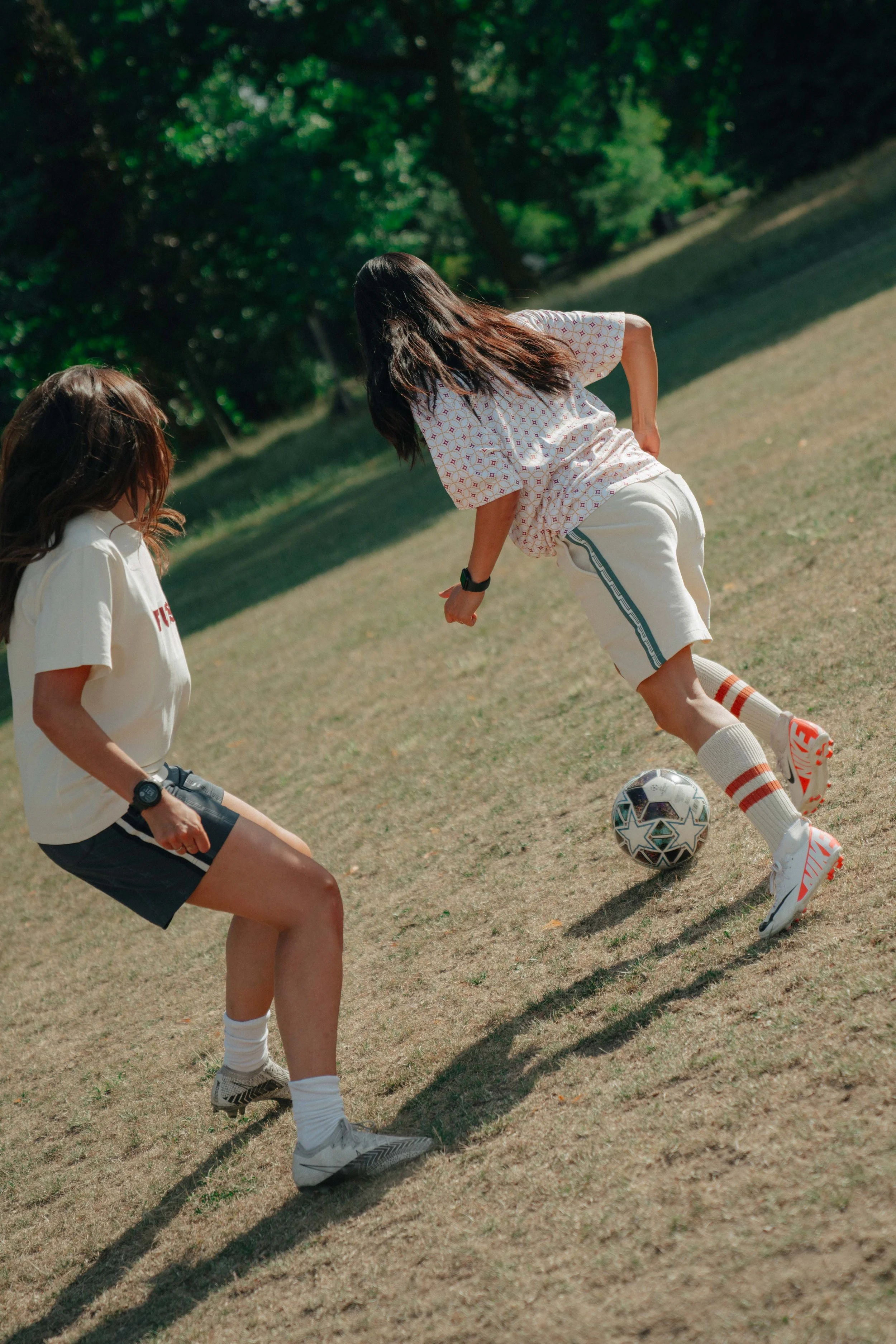 Two women playing soccer outdoors on a grassy field surrounded by trees.