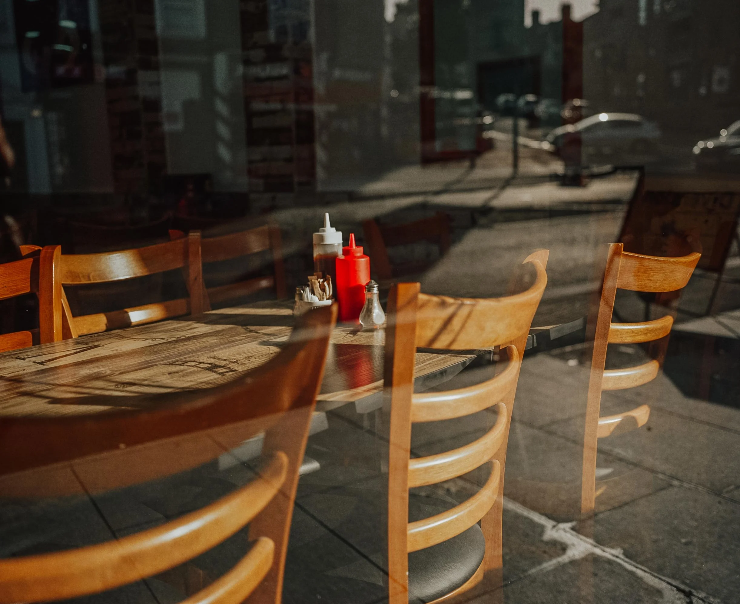Empty outdoor dining area with wooden chairs and table, condiments, and utensils, viewed through a glass window with reflections of parked cars outside.