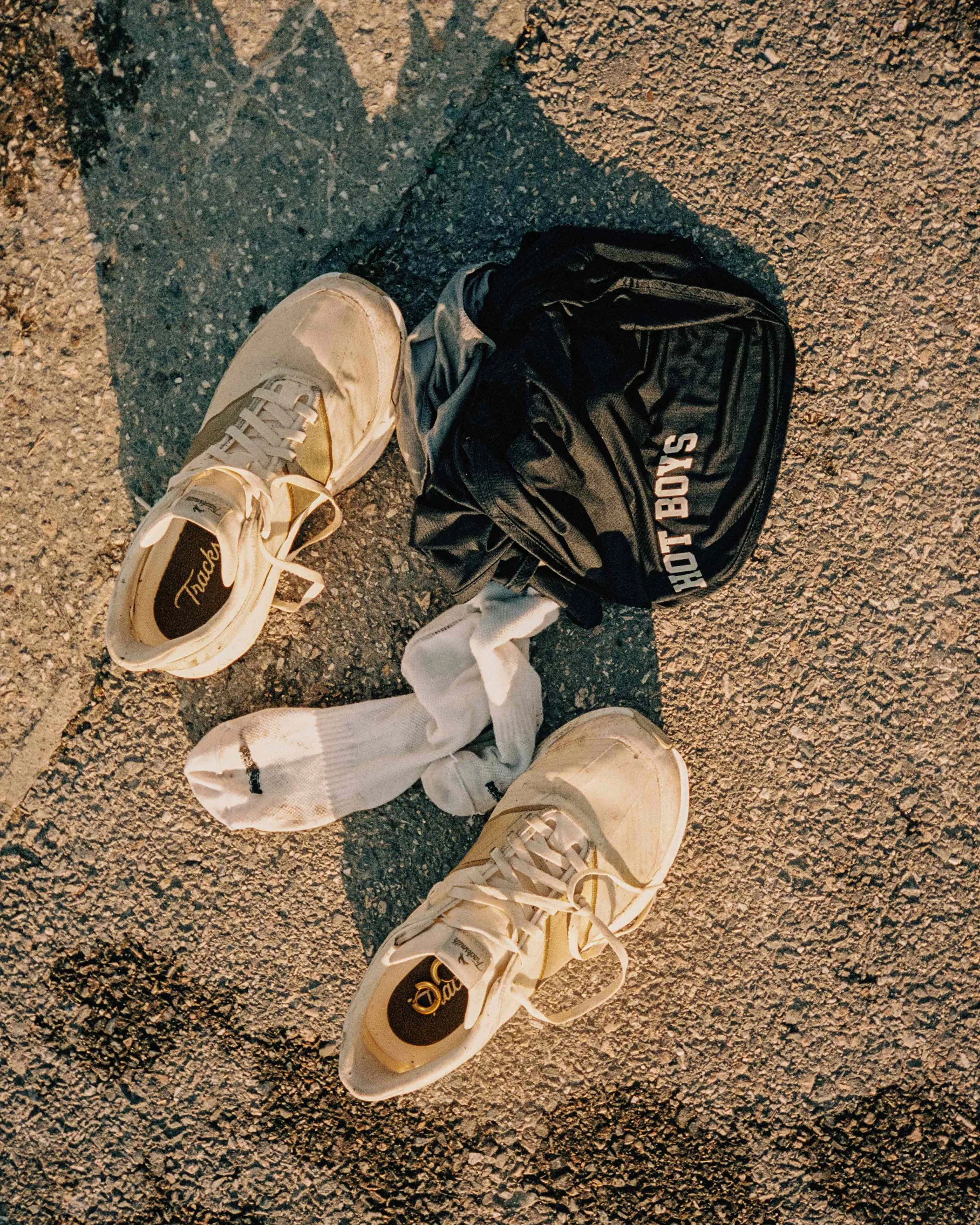 A pair of white sneakers, white socks, a black backpack labeled 'HOT BOYS', and a white shirt are on a rough-textured pavement, illuminated by warm light.