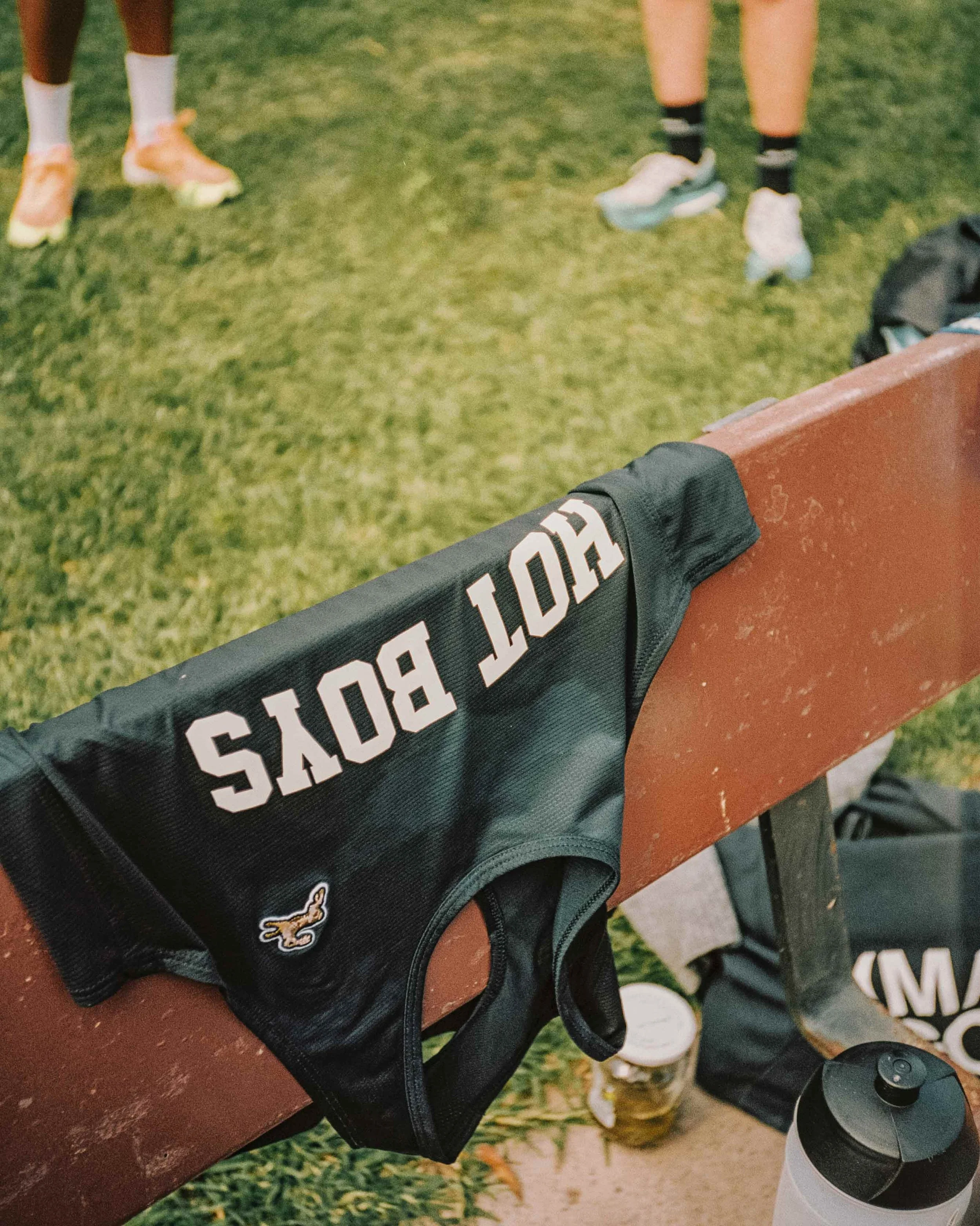 A black sports jersey with white text reading 'HOT BOYS' draped over a park bench, with a grassy field and people in athletic clothing in the background.