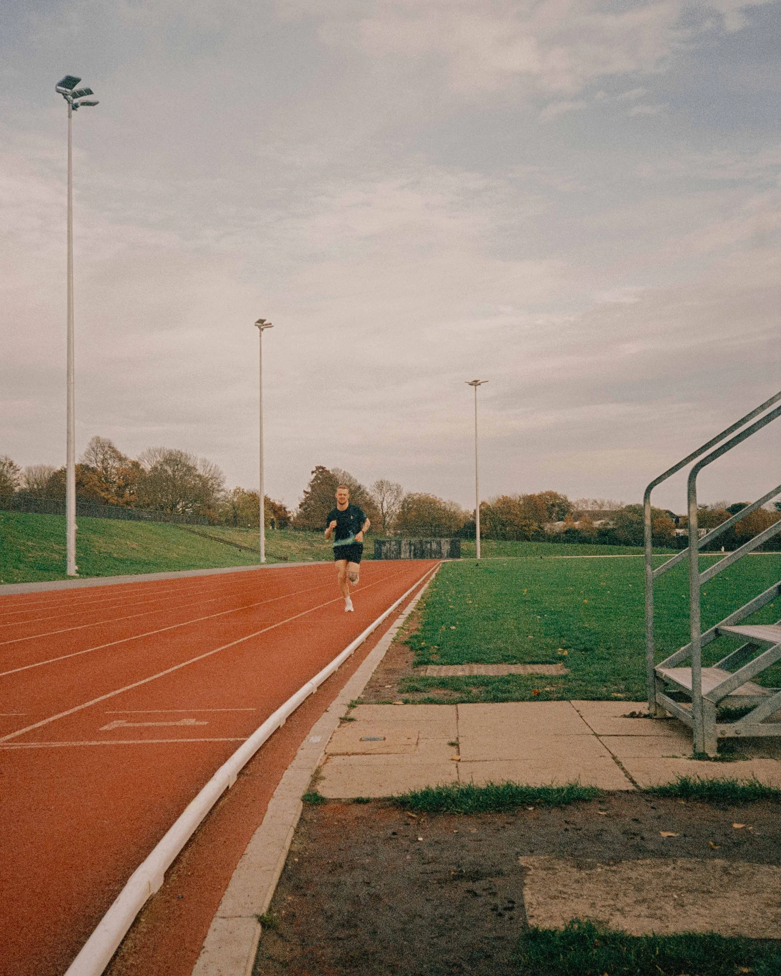 A man running on a red outdoor track with green grass on the sides, under a cloudy sky, with light poles and trees in the background.