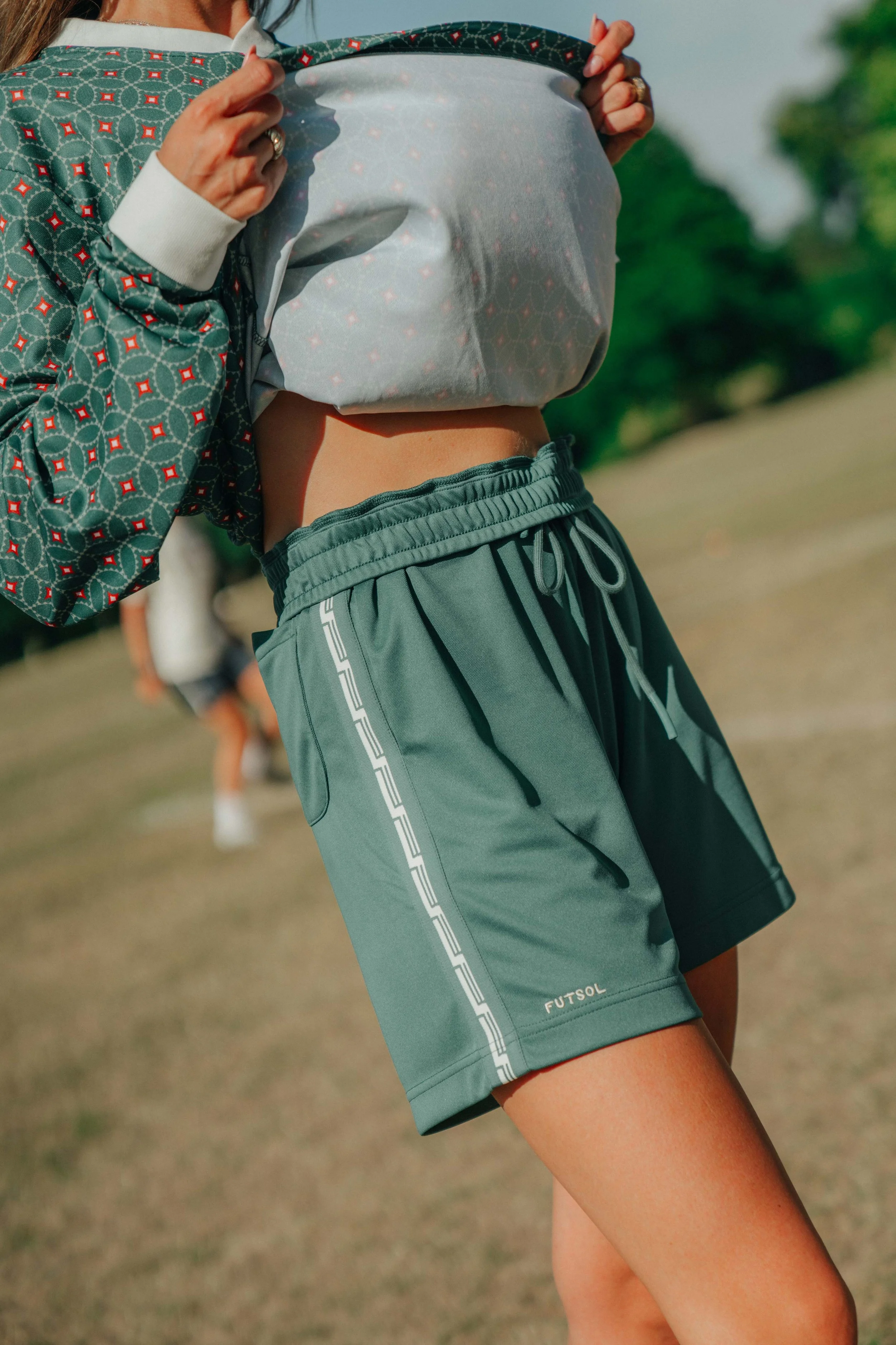Person lifting a patterned jacket to reveal a crop top, wearing green Futsal shorts, outdoors on a grassy field.