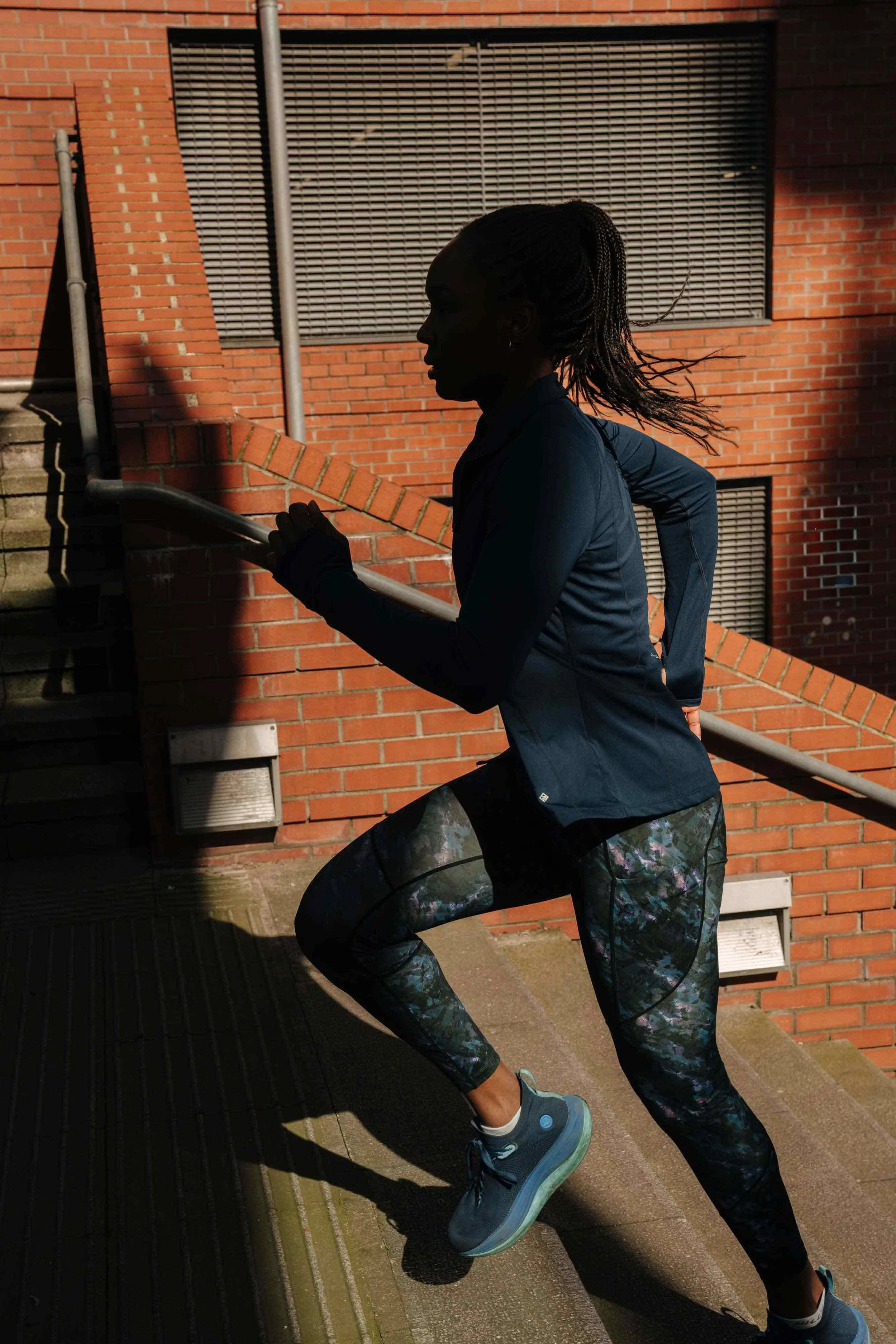 A woman running on outdoor stairs, wearing athletic clothing, viewed in silhouette with brick building background.