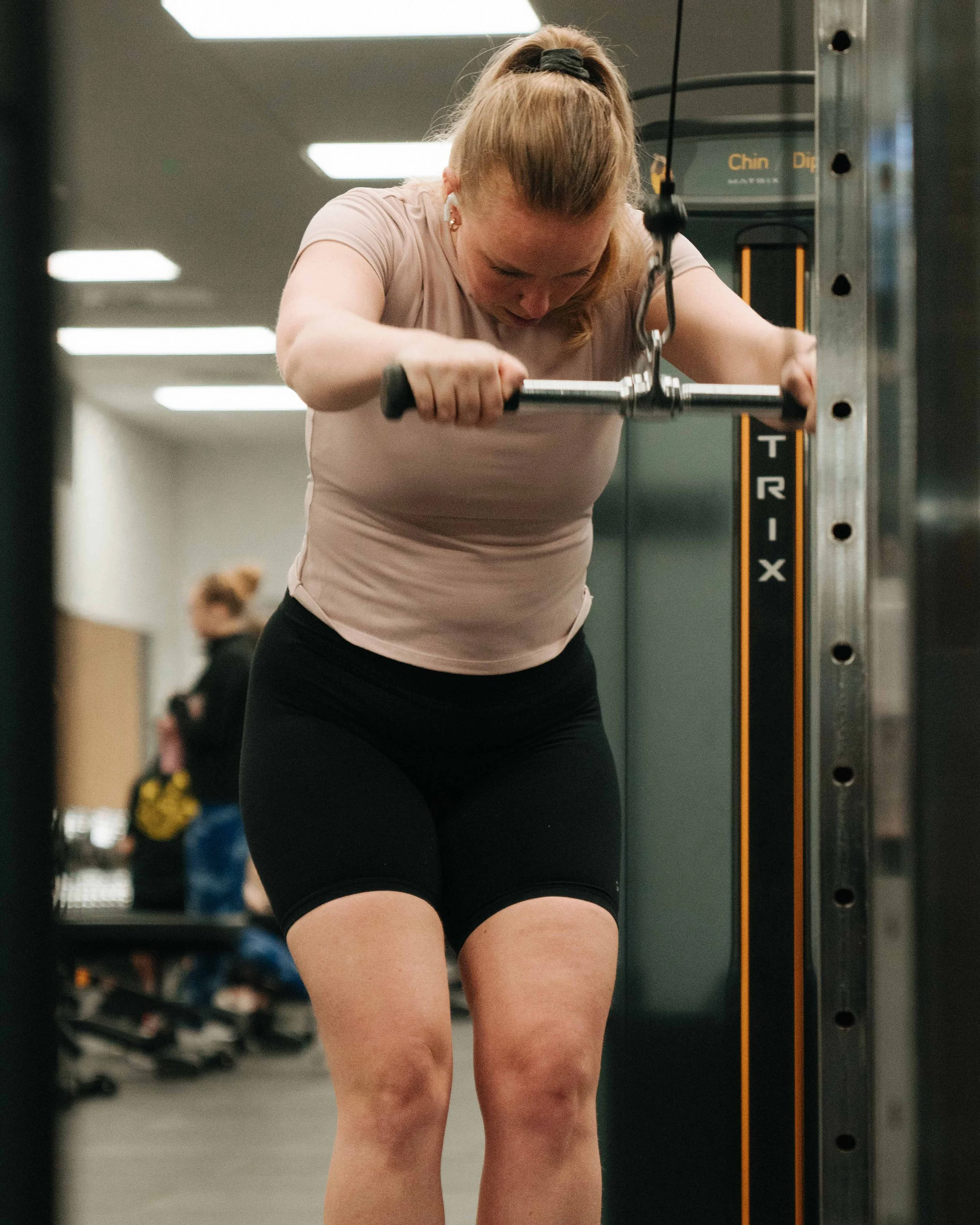 A woman with red hair tied back in a ponytail is doing an exercise, stretching her back using a TRX suspension trainer in a gym. She is wearing a light pink t-shirt and black shorts. In the background, there is another person and gym equipment.