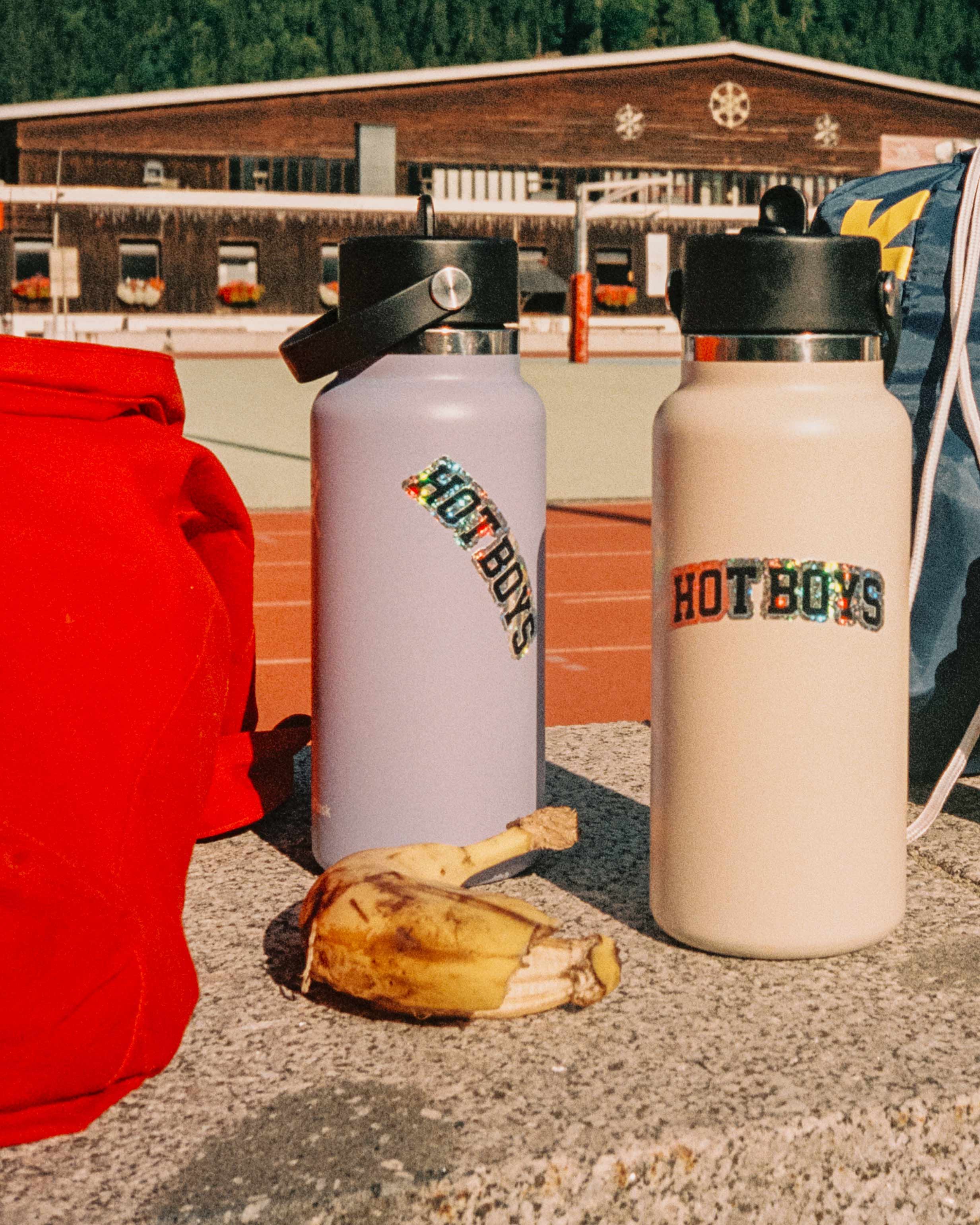 Two water bottles labeled 'Hot Boys' and 'Hot Boys' with a banana peel in front, set on a concrete surface outdoors with a red backpack, a blue bag, and a building in the background.