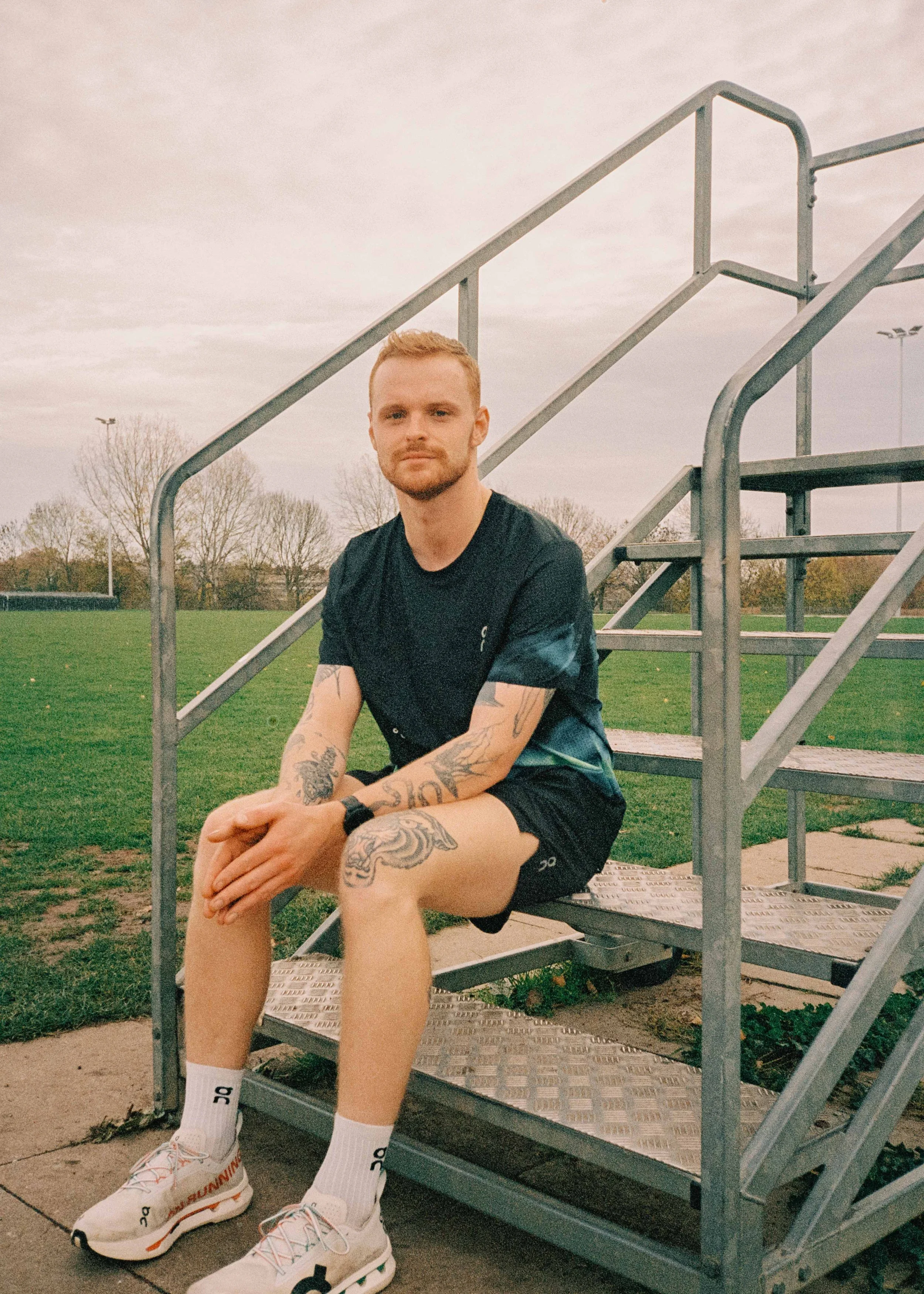 A young man with tattoos sitting on metal stairs at a sports field, wearing black athletic shorts, a black t-shirt, white sneakers, and white socks.