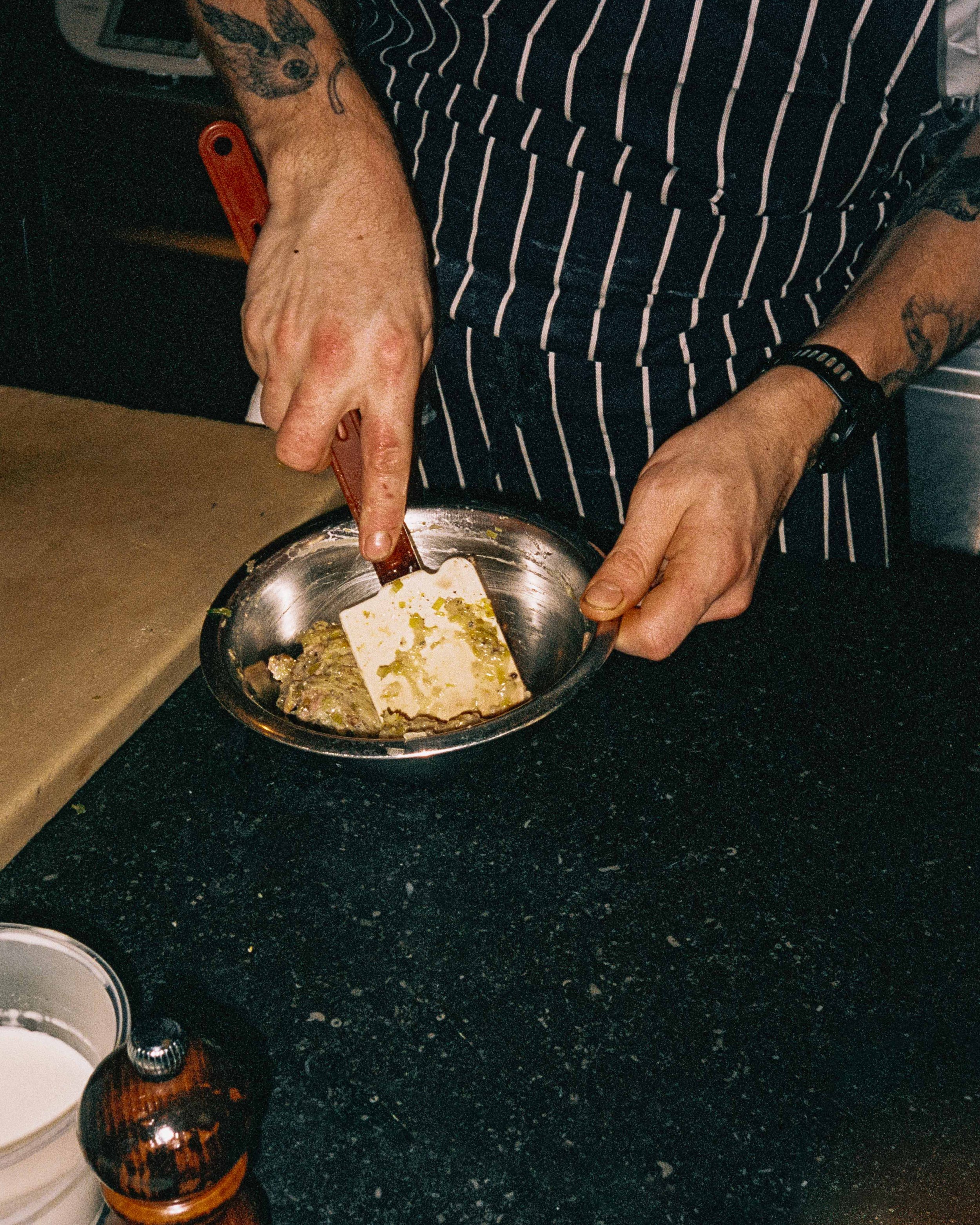 Person wearing a striped apron or shirt, cooking or preparing food in a kitchen, using a spatula to stir or scrape a dish in a small stainless steel bowl.