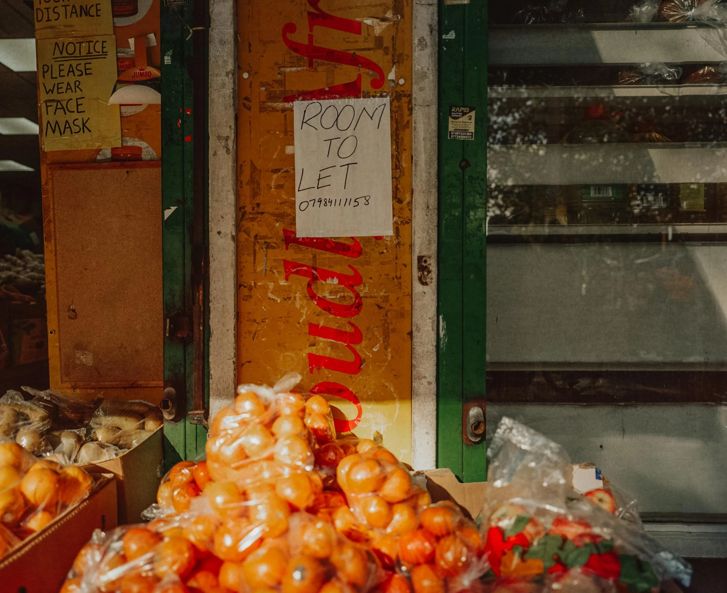 A small grocery store display with onions in plastic bags in the foreground, a sign that reads 'Room to Let' with a phone number, and a notice asking customers to wear face masks.