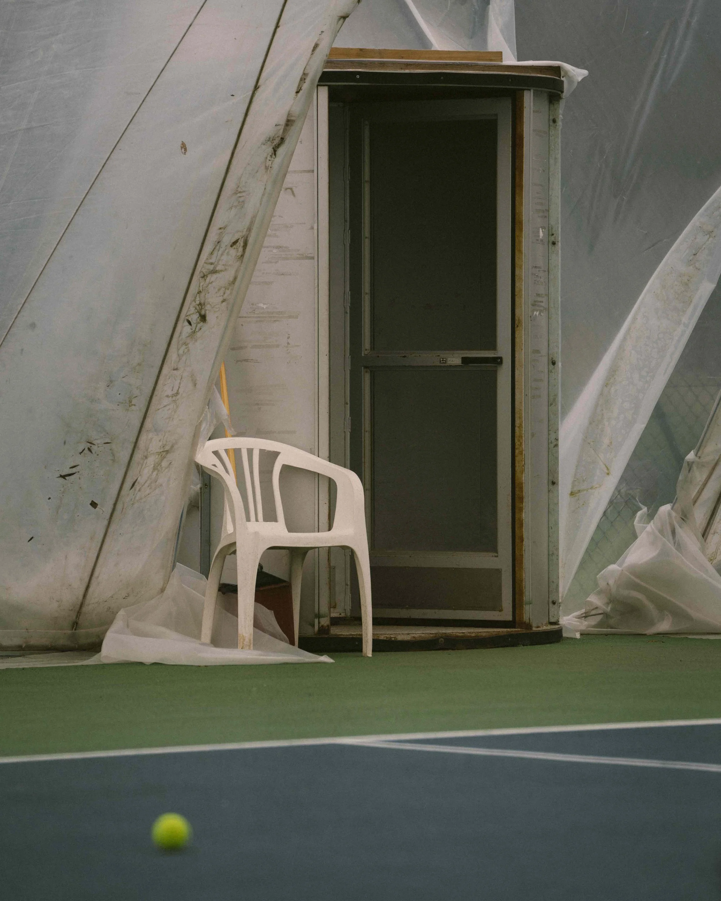 An indoor tennis court with a white plastic chair, a tennis ball on the court, and a mirror or glass panel with a metal frame leaning against an unfinished wall.