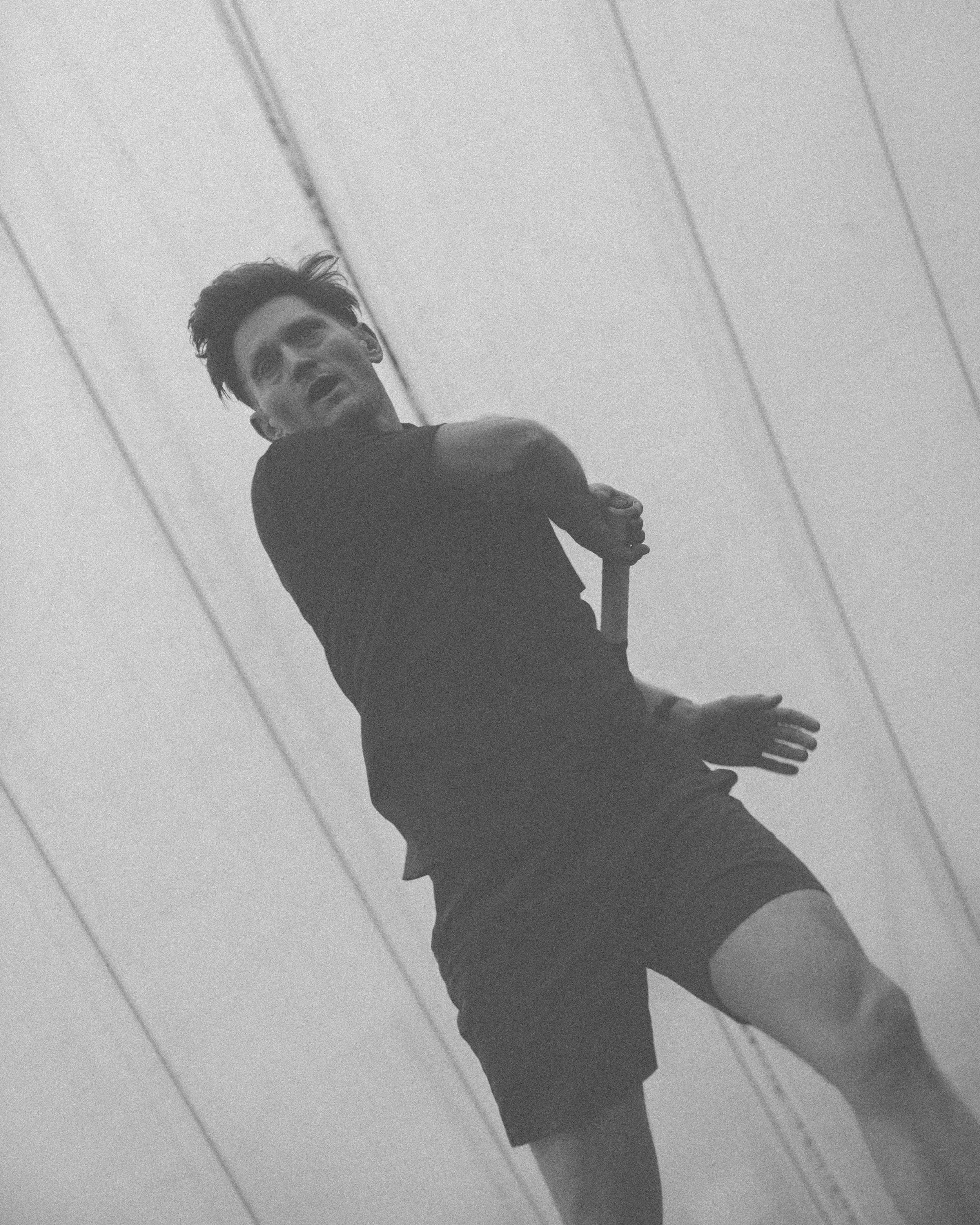 A black and white photo of a young man with dark hair, wearing a dark long-sleeve shirt and shorts, standing in front of a mirrored wall, looking sideways with a serious expression.