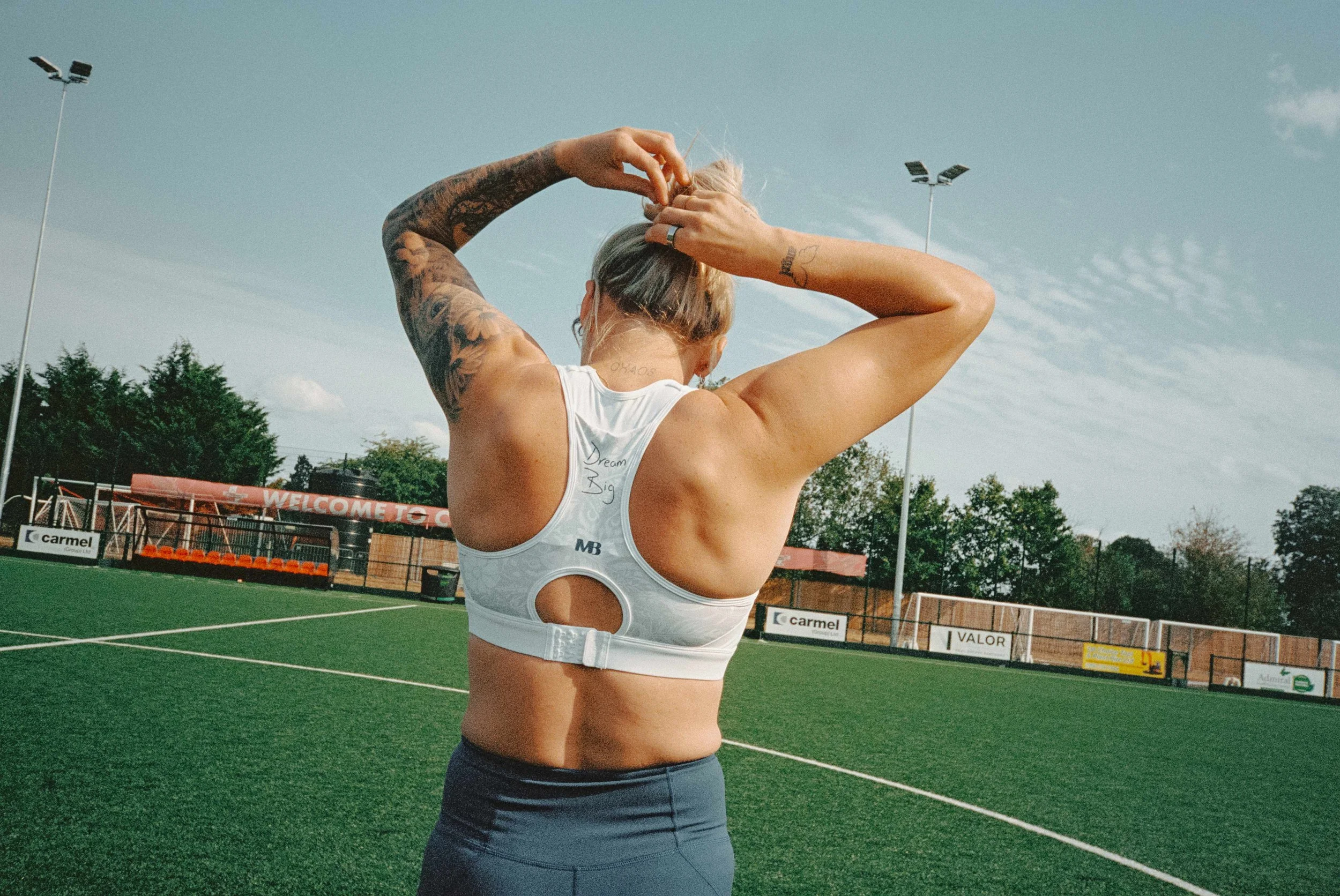 A woman with tattoos on her arms, wearing a white sports bra and dark athletic leggings, standing on a green sports field, seen from behind as she adjusts her blonde hair.