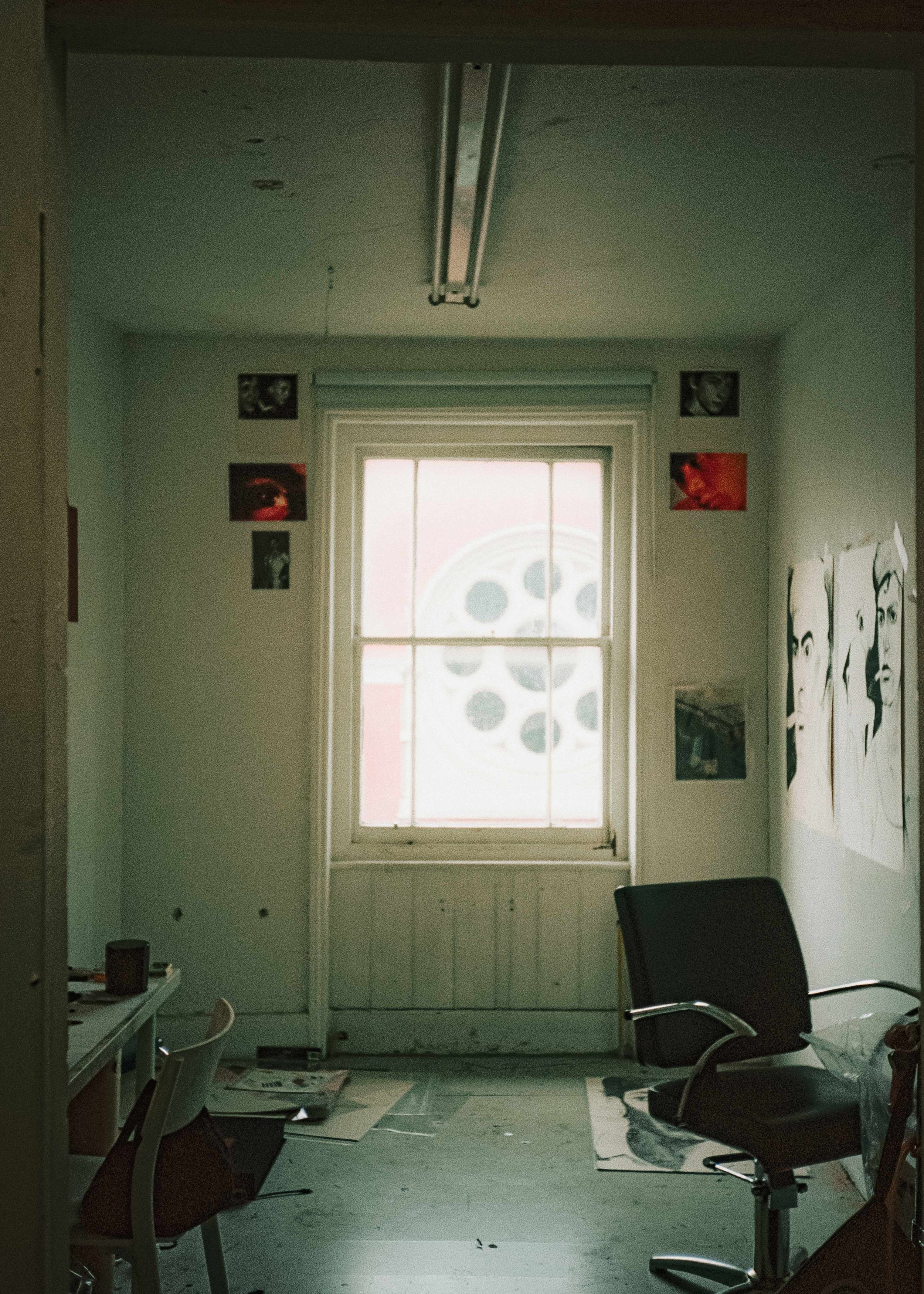 A room with a window with frosted glass featuring a circular pattern, surrounded by small collage of photos on walls, a black office chair, and a desk on the left with scattered items.