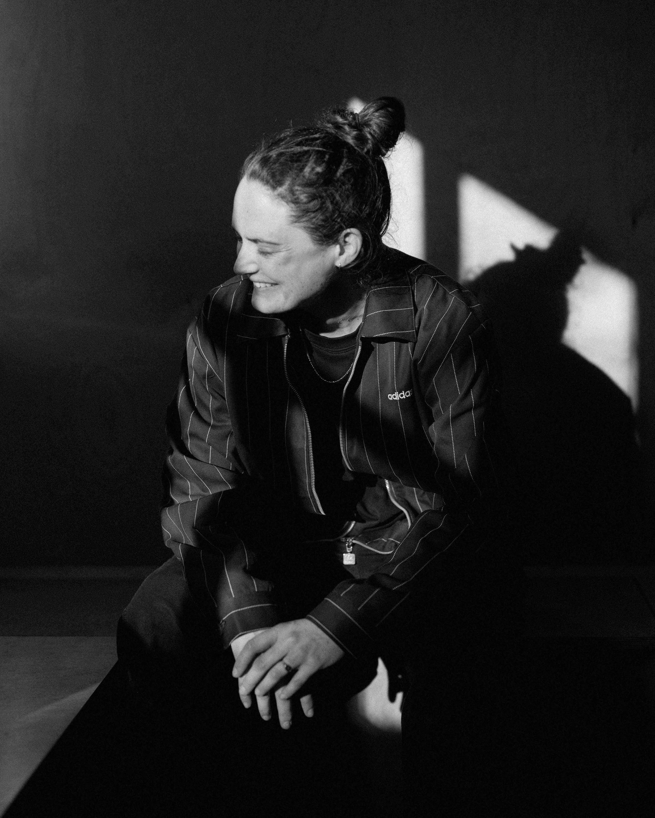 Black and white photo of a woman smiling, sitting with her hands clasped, leaning slightly forward, with a shadow cast on the background.