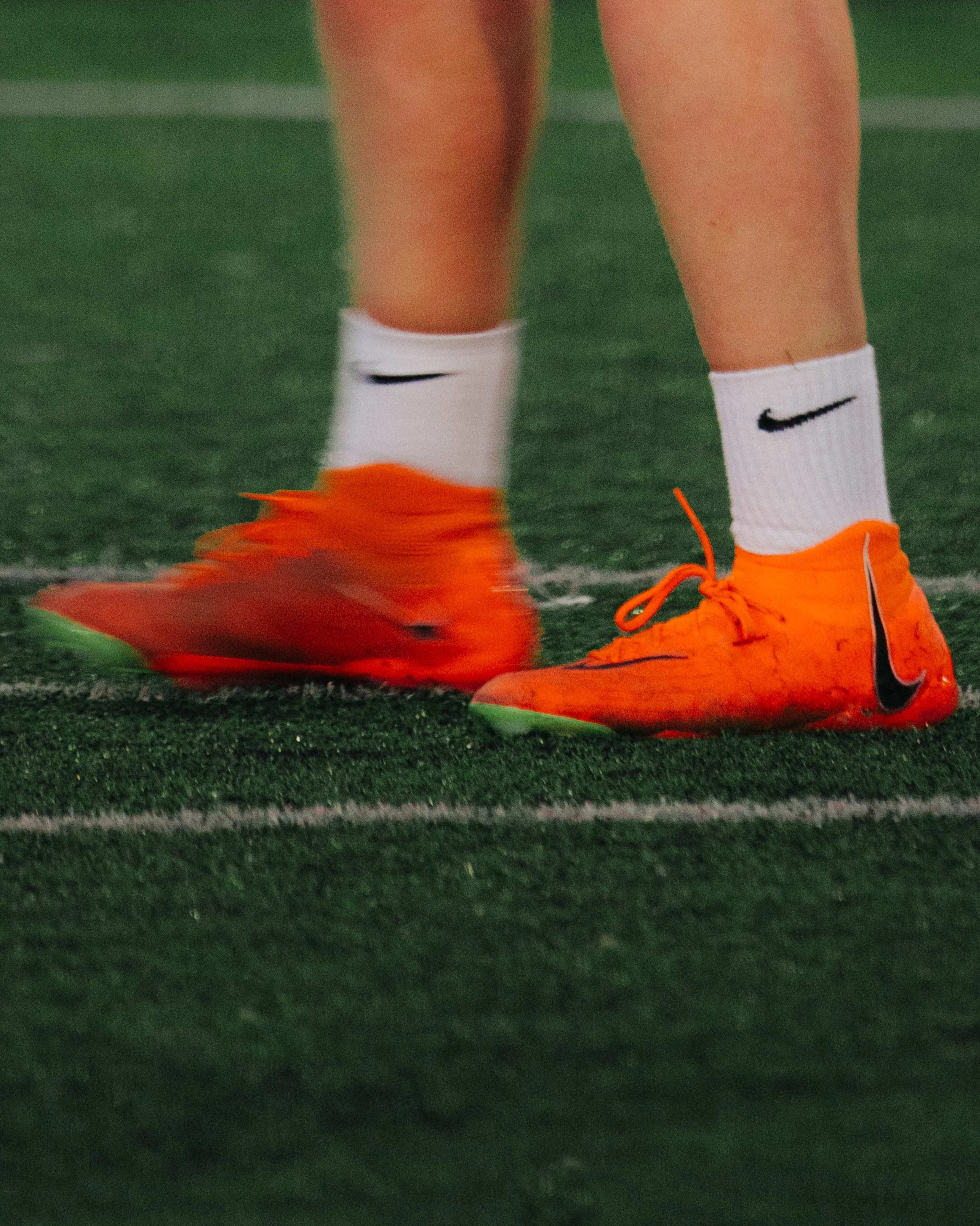 Close-up of a soccer player's feet wearing bright orange Nike football shoes and white Nike socks on a green turf field.